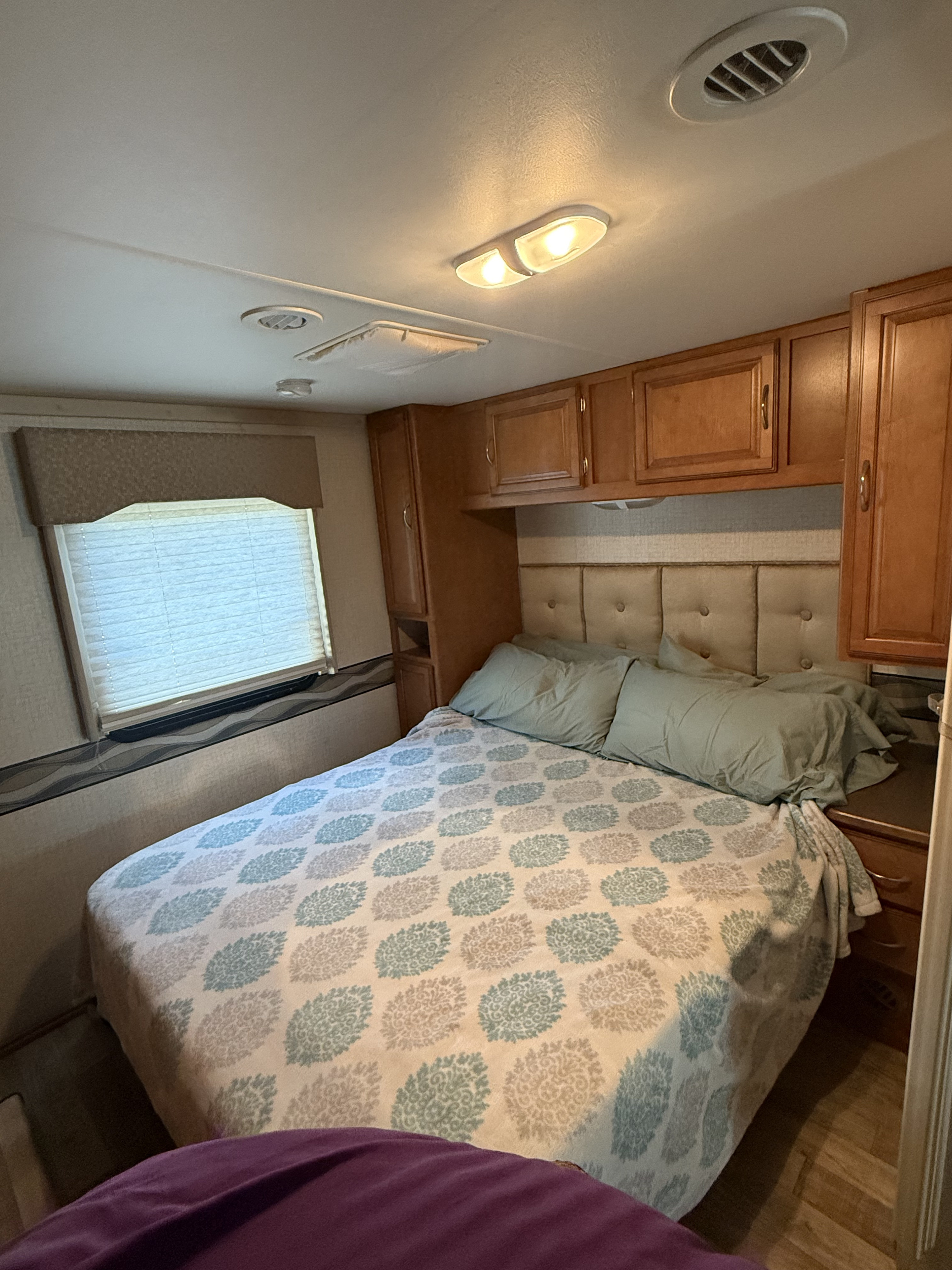 RV rental bedroom with a comfortable bed, patterned bedding, window, and light wooden cabinets for storage.. Winnebago Minnie Winnie 2017