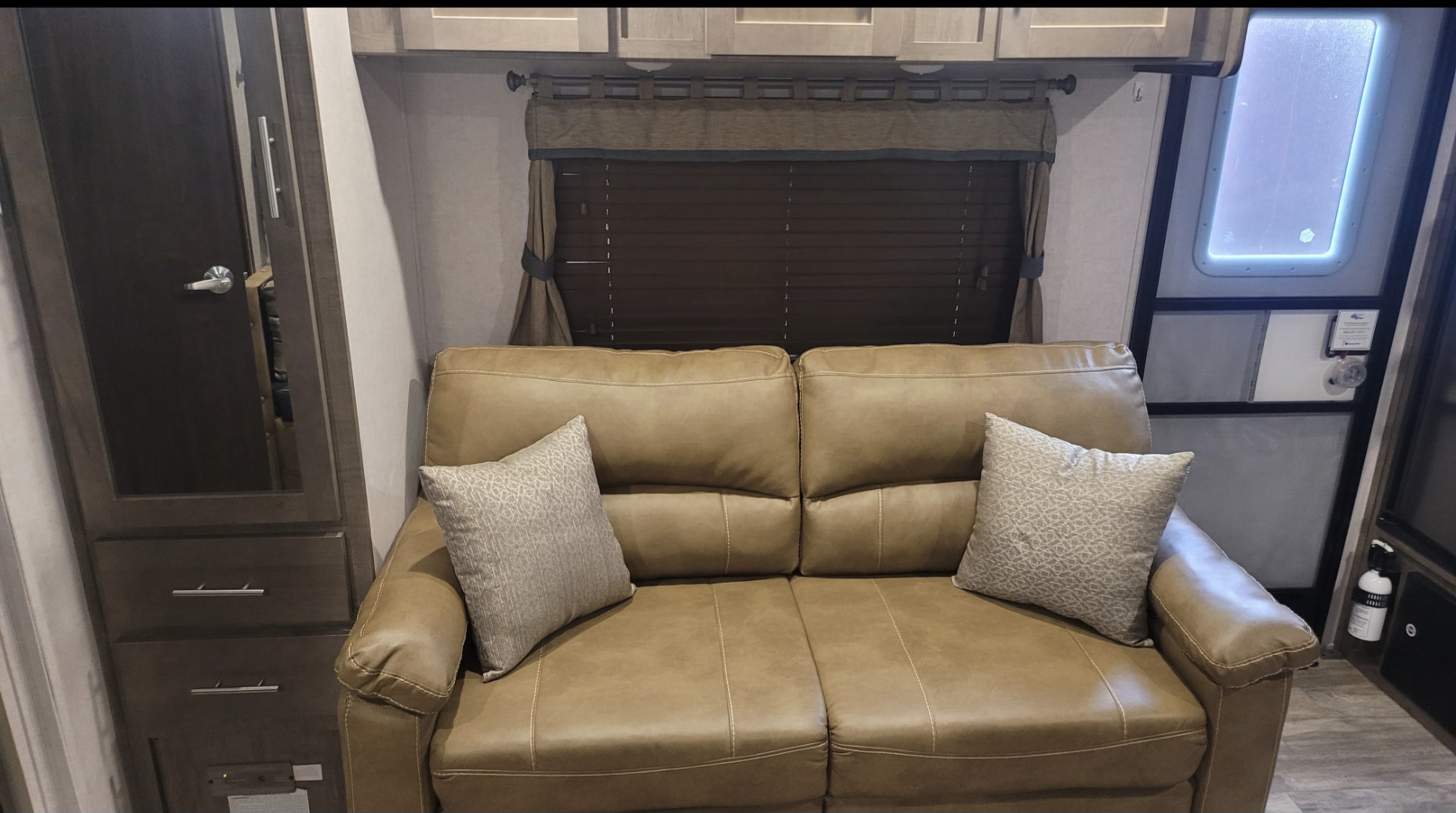Cozy RV rental living area featuring a tan sofa with pillows, window with blinds, and storage. Fire extinguisher by door.. Forest River Surveyor Legend 2021