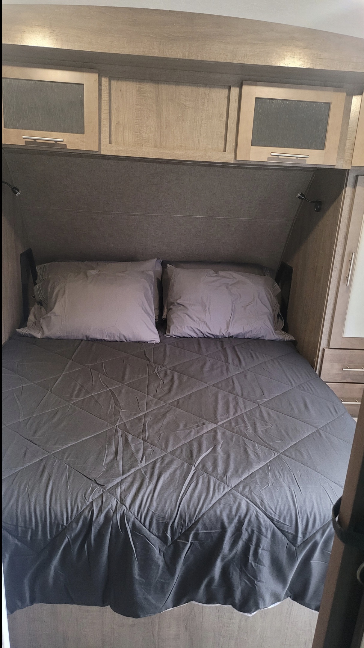 Well-appointed bedroom in an RV rental, showing a gray bed with quilted comforter, pillows, and wood-paneled overhead cabinets.. Forest River Surveyor Legend 2021