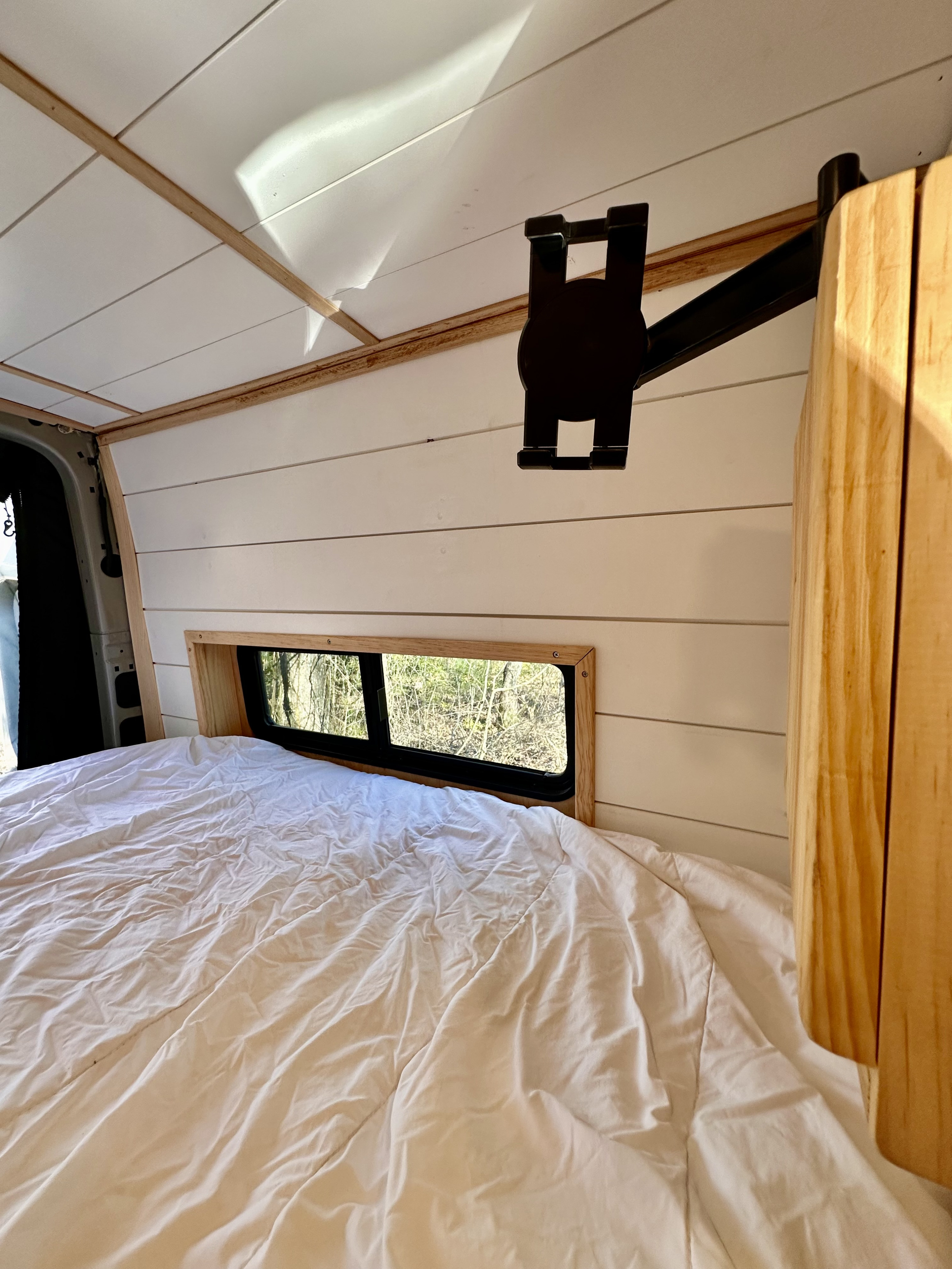 This RV rental's cozy sleeping area features a comfortable bed, white paneled walls, and a window with a tree view.. Mercedes-Benz Sprinter 2022