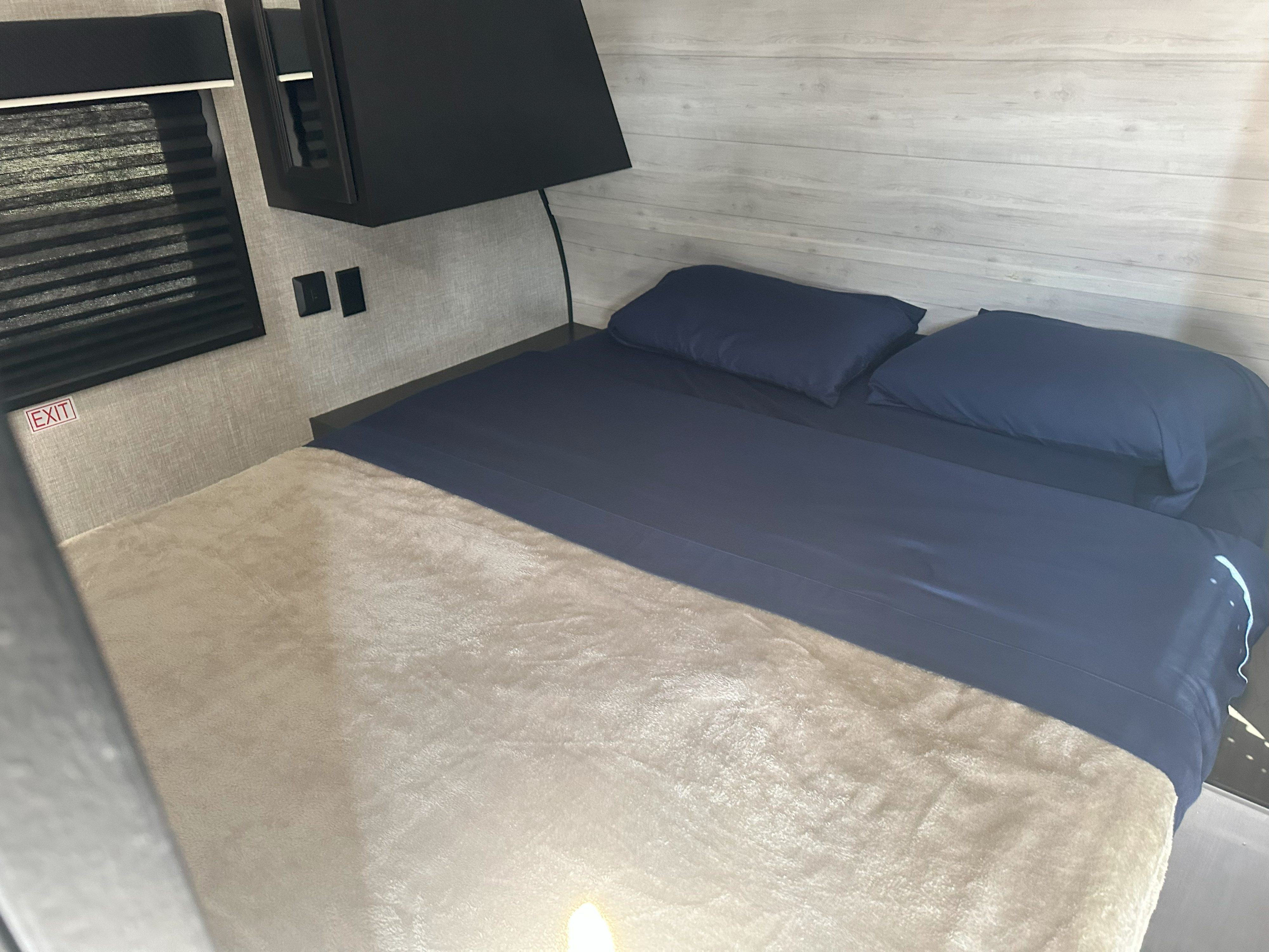 Cozy rv rental bedroom showing a compact bed with dark blue linens and a light blanket. Window and 'EXIT' sign on wall.. Jayco Jay Flight 2022