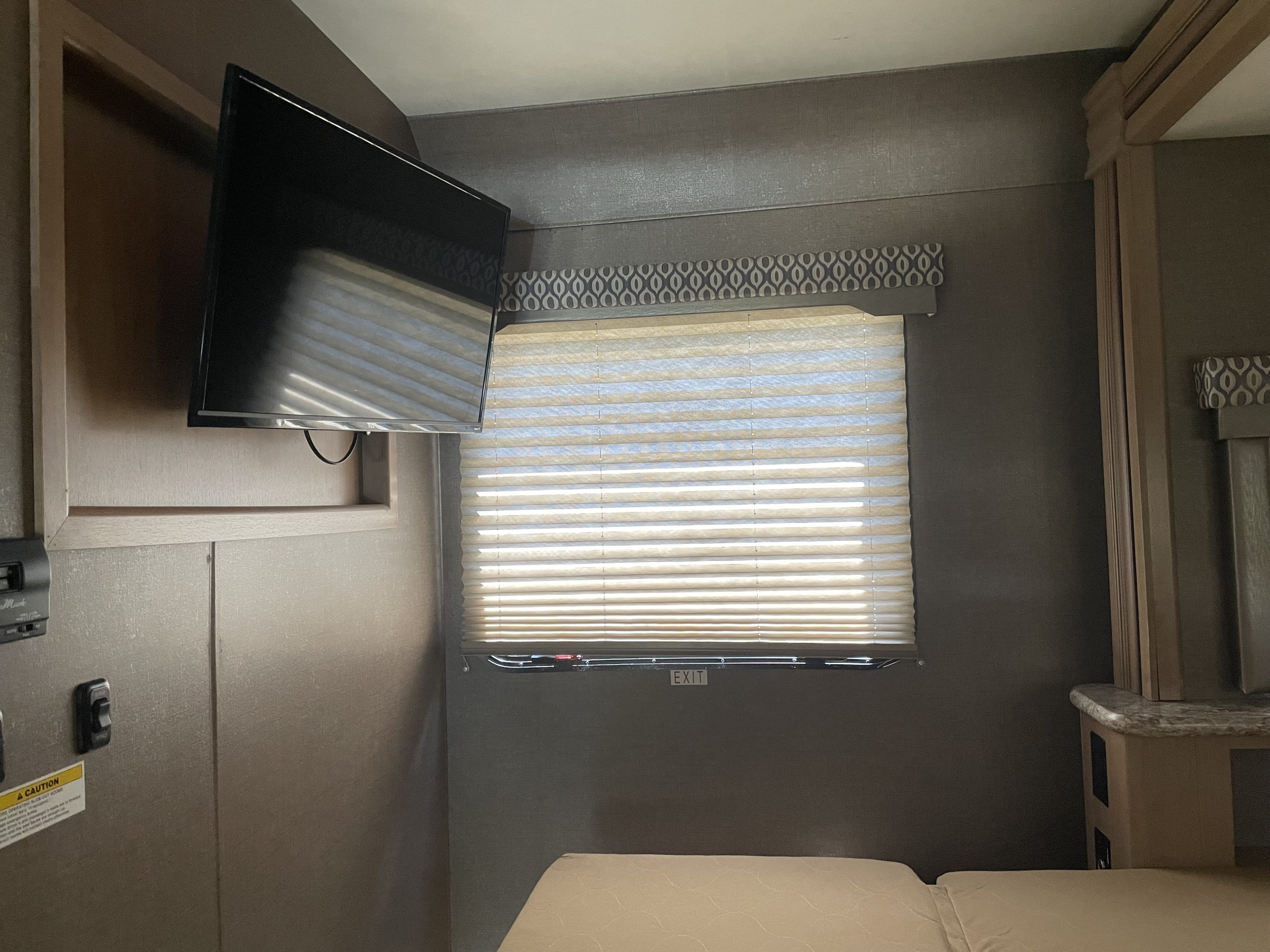 View inside an RV rental bedroom showing a wall-mounted TV, window with pleated blinds, and a bed. Includes an emergency exit.. Thor Motor Coach Freedom Elite 2019