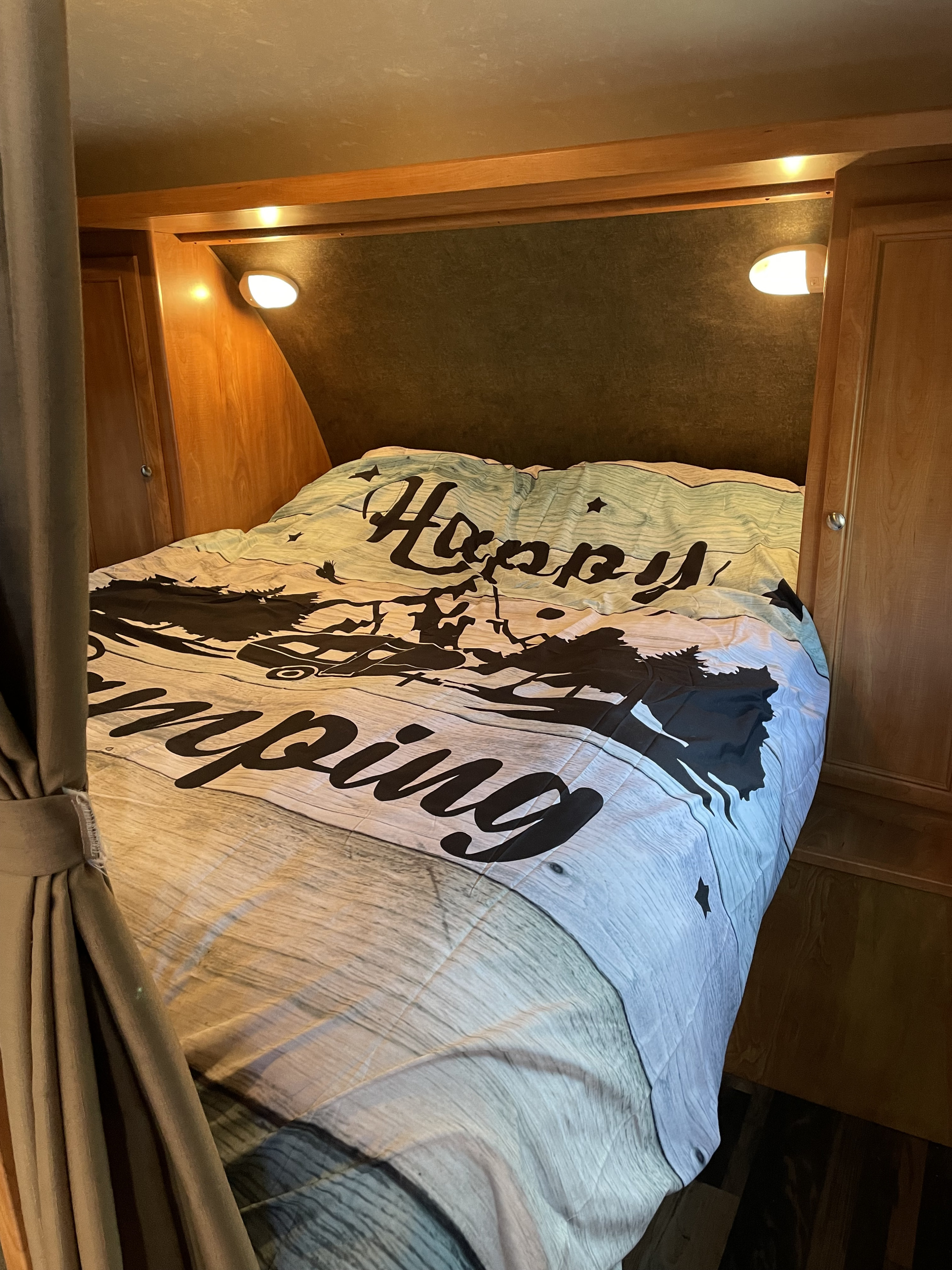 Cozy RV rental bedroom with "Happy Camping" comforter. Features wooden cabinets, lighting, and a curtain.. Forest River Shasta Flyte 2015