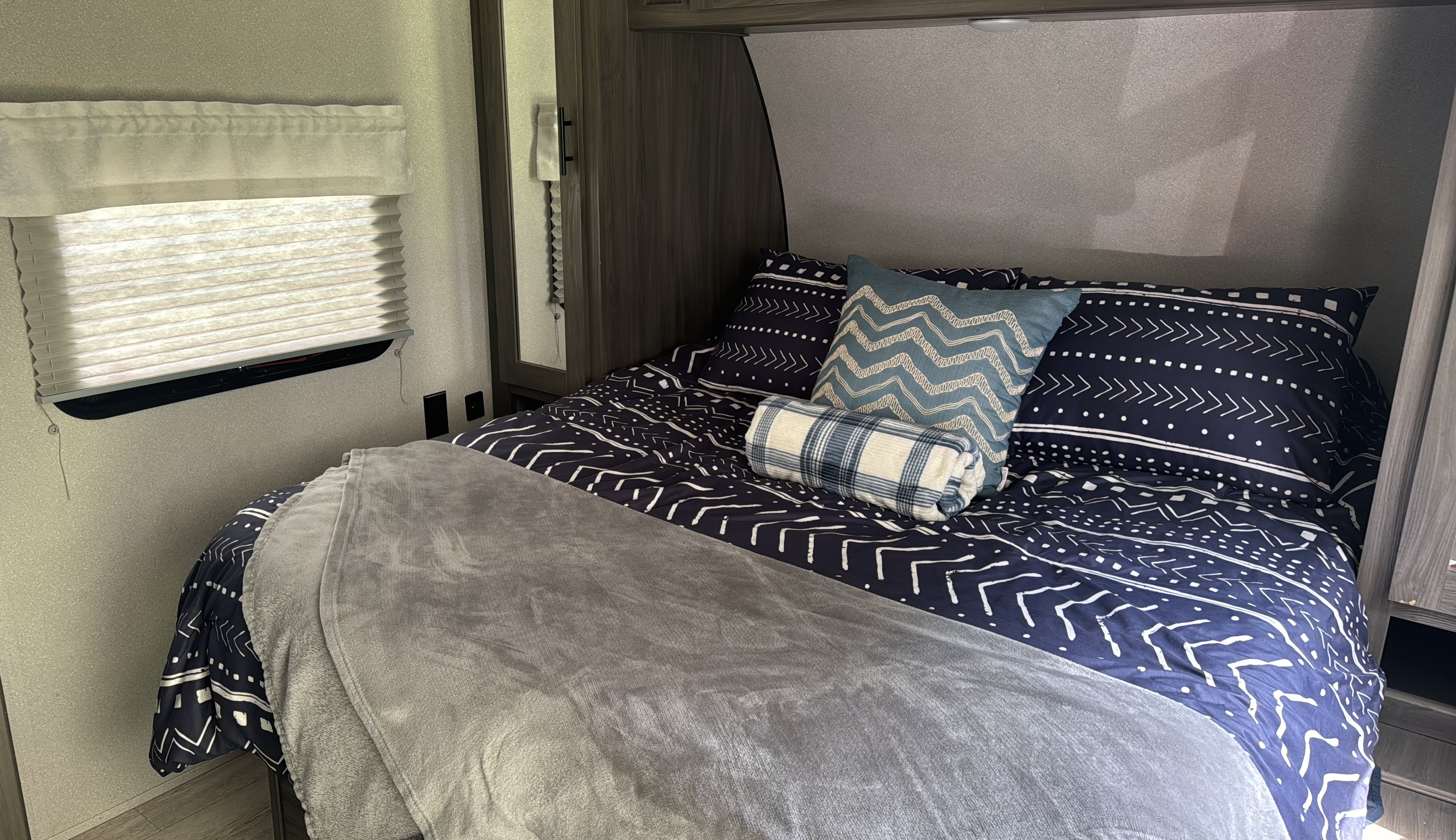 Cozy RV rental bedroom featuring a bed with blue patterned bedding, a light blue zigzag pillow, and a grey blanket for comfortable rest.. Forest River Shasta 2021