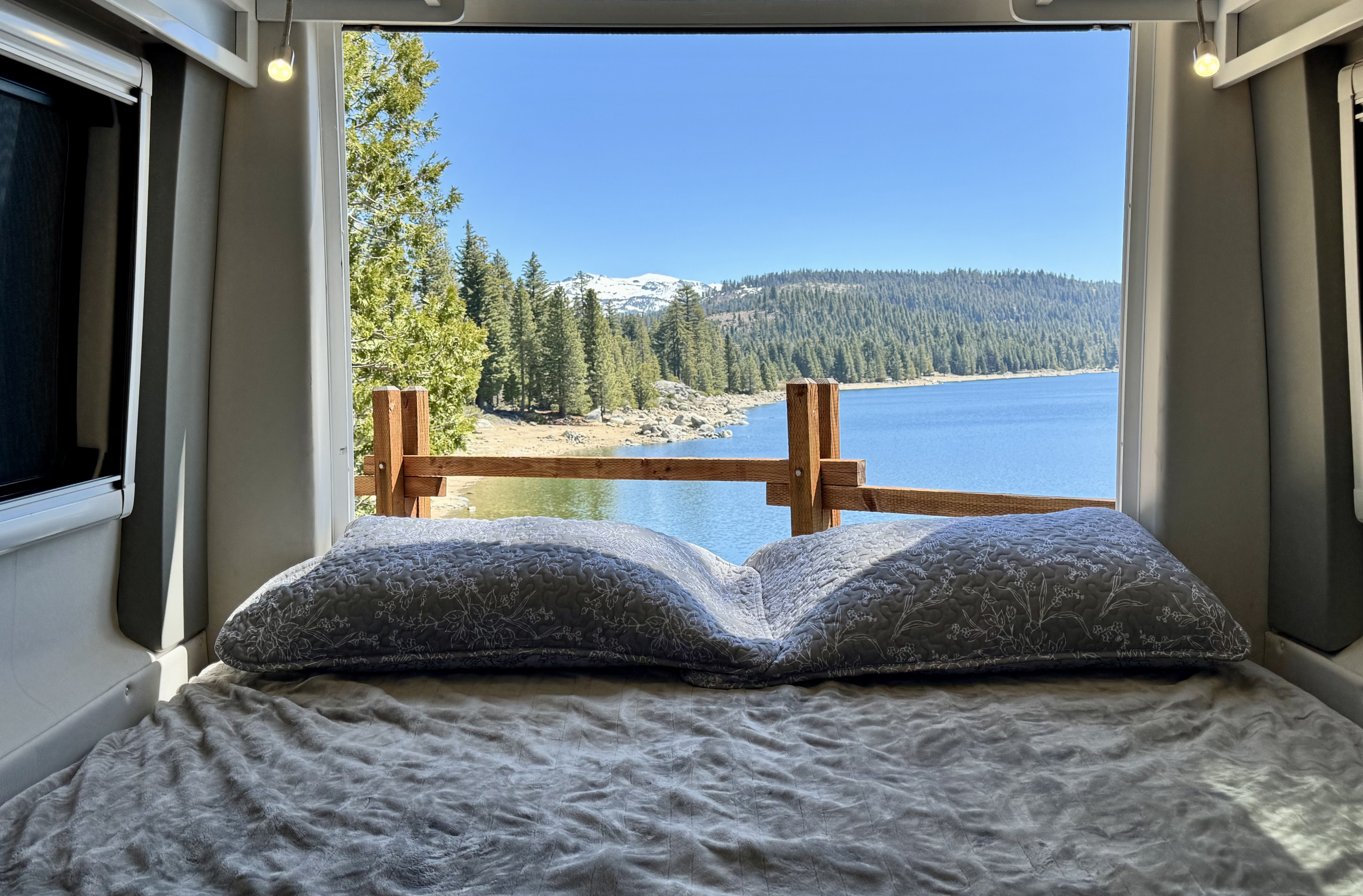 Inside an RV rental, a cozy bed faces a panoramic view of a tranquil lake, lush forest, and distant snow-capped mountains.. Coachmen Beyond 2021