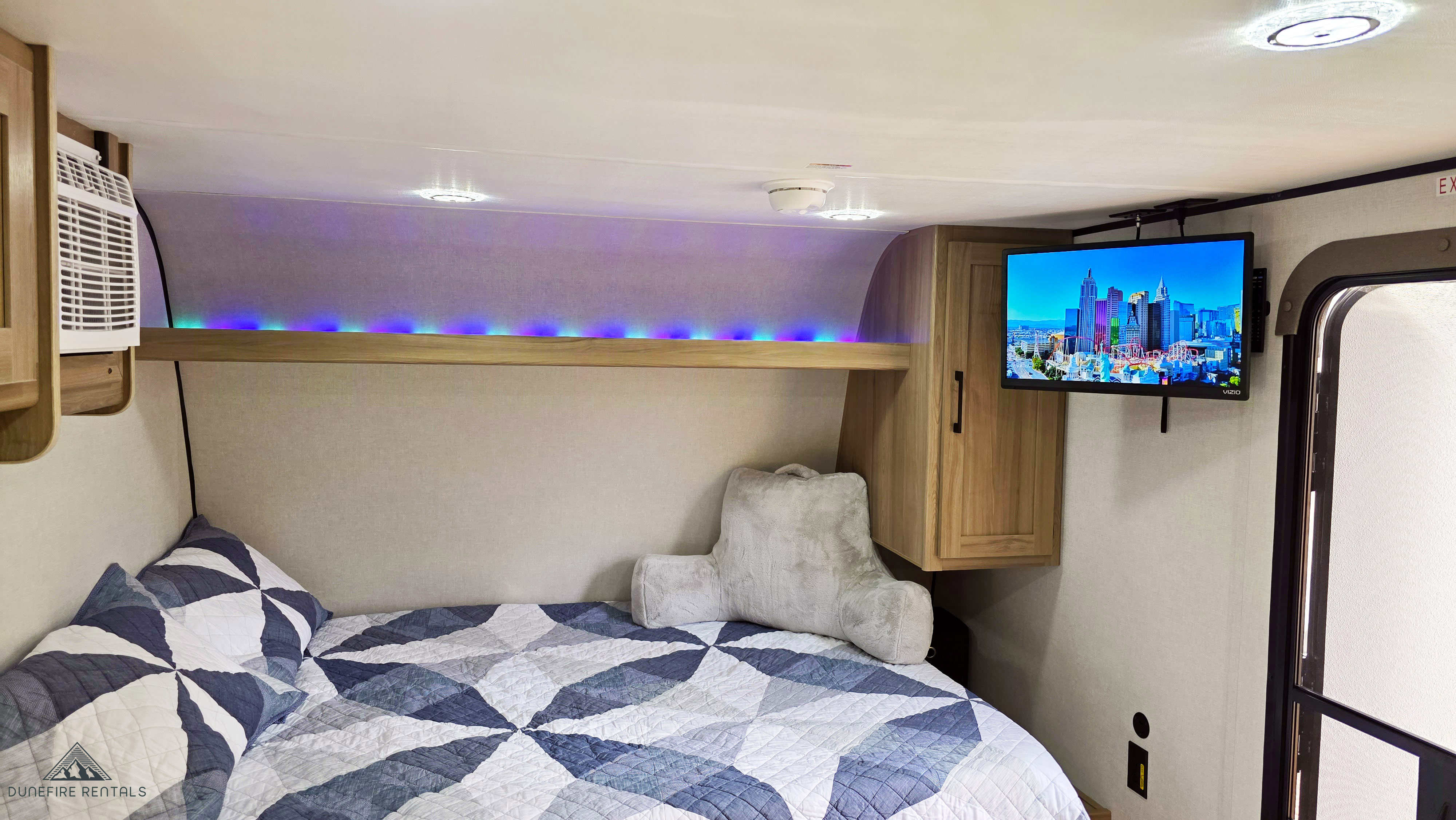 Cozy rv rental bedroom showing a bed with a blue/white quilt, colorful LED lights, a mounted TV, and wood cabinets.. Keystone Coleman 17B 2025