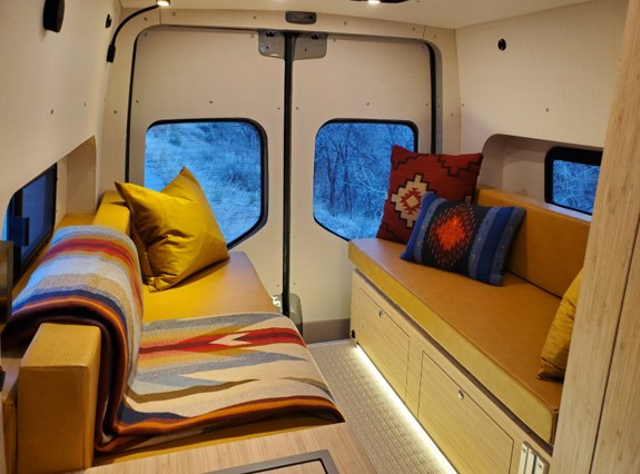 Cozy RV rental interior featuring yellow bench seating with colorful throws and pillows, offering scenic winter forest views.. Mercedes Sprinter 2021