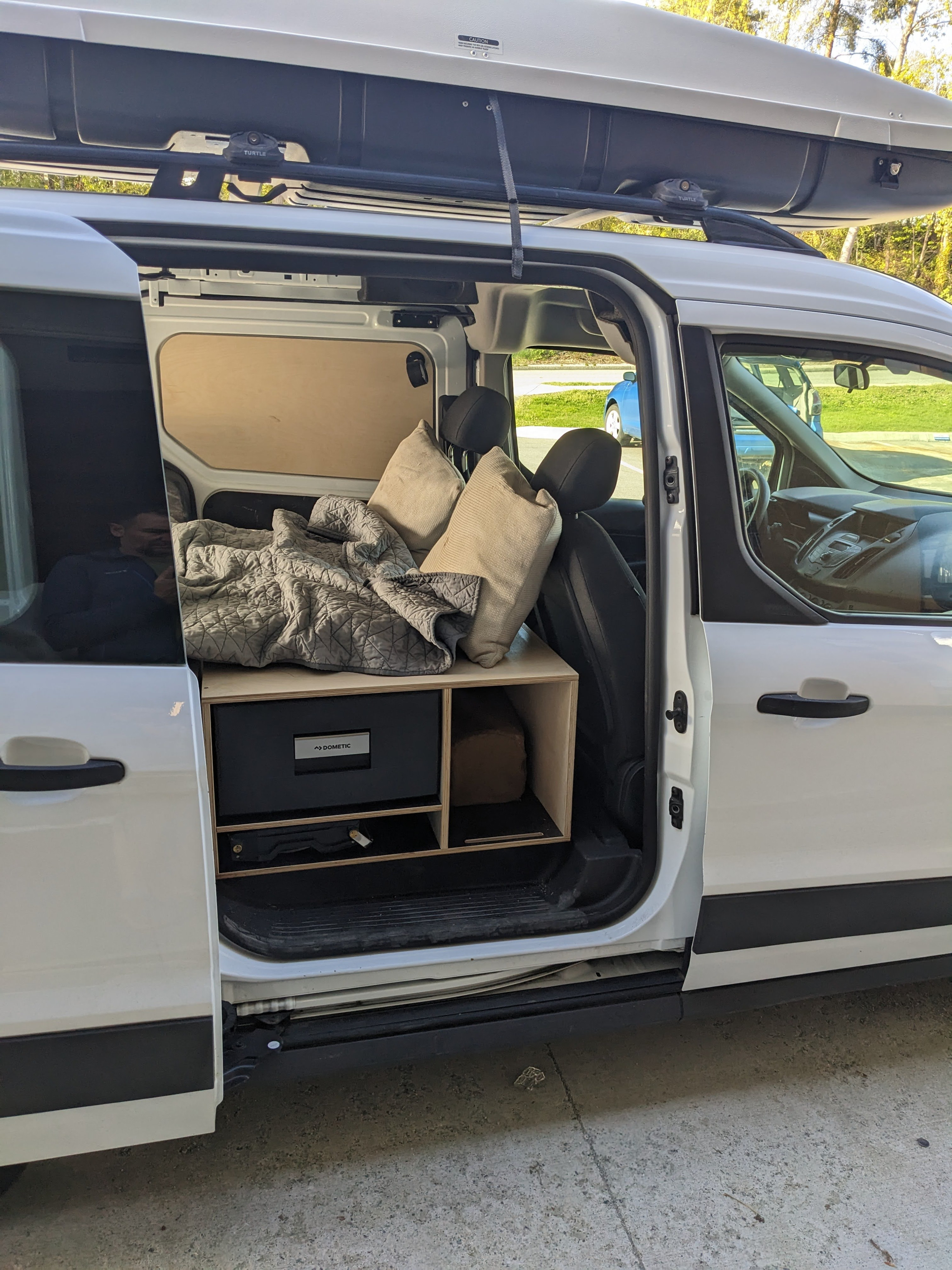 White camper van rv rental. Open door shows custom interior with bed, pillows, Dometic fridge, and storage shelves.. Ford Transit 2015