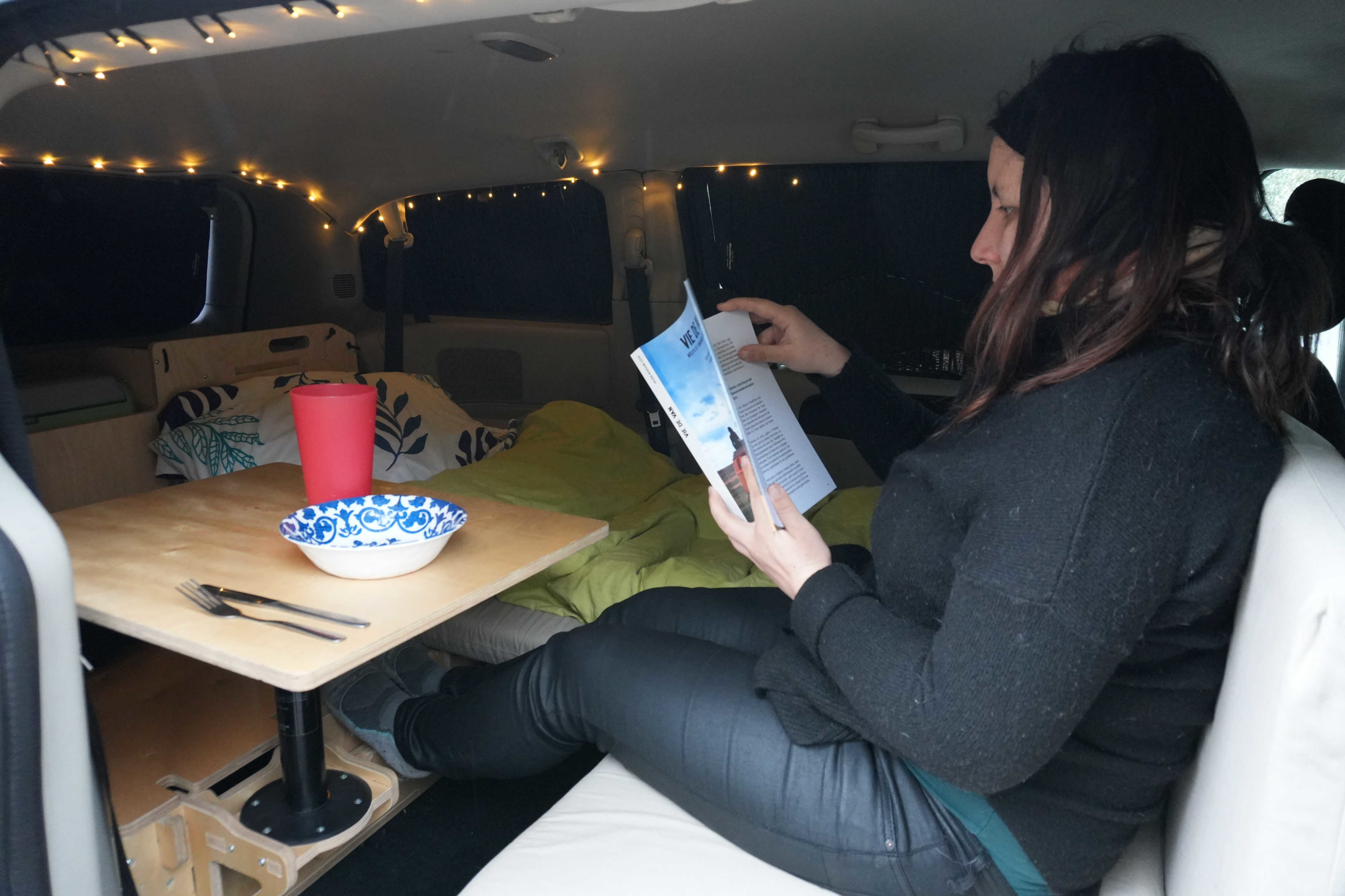 A person relaxes inside a cozy, customized RV rental, reading a book at a small table, illuminated by string lights.. Dodge Grand Caravan 2014