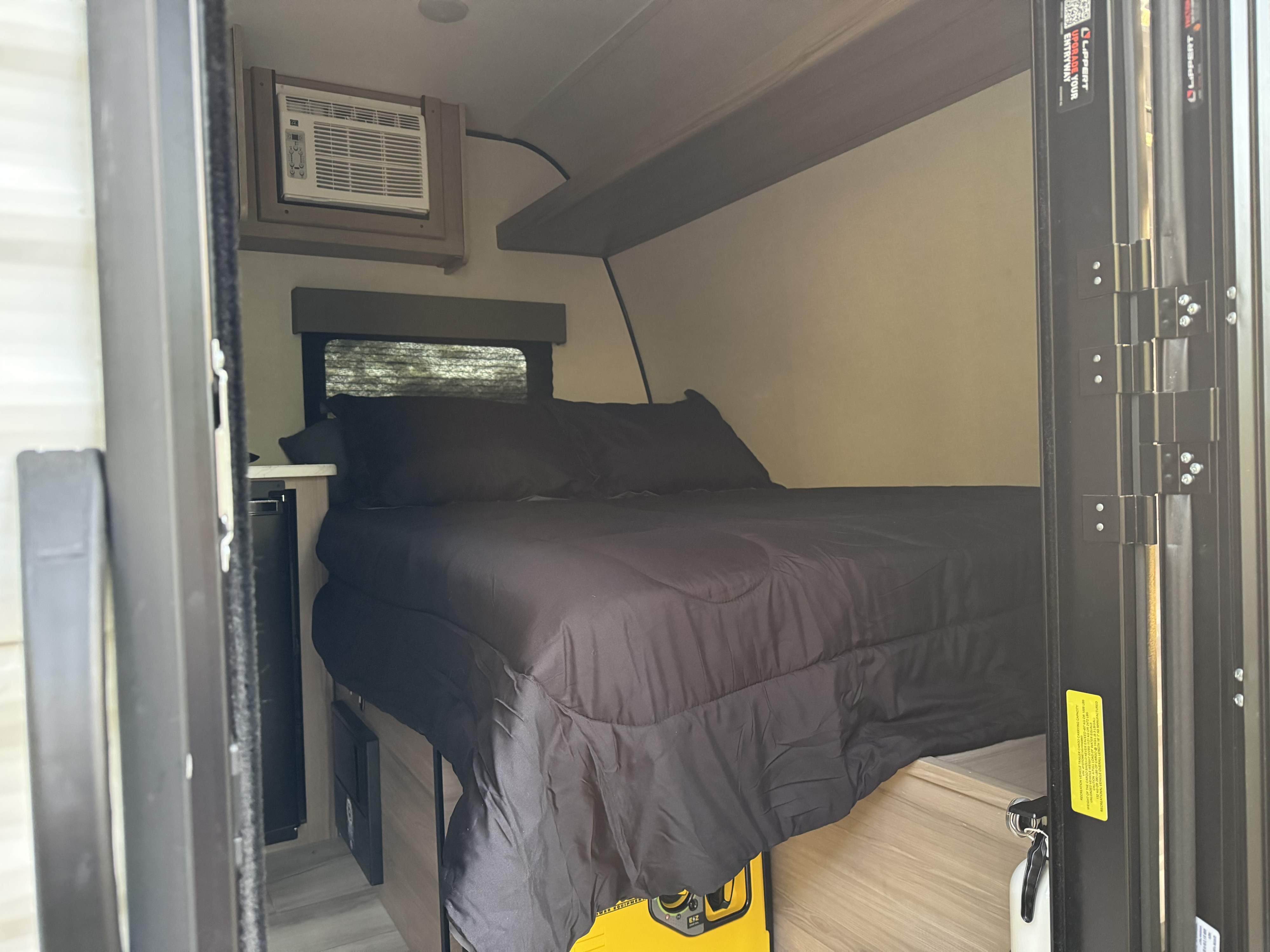 Cozy RV rental bedroom with black bedding, AC unit, window, and portable power. Compact and comfortable for travel.. Keystone RV Coleman 13B 2025