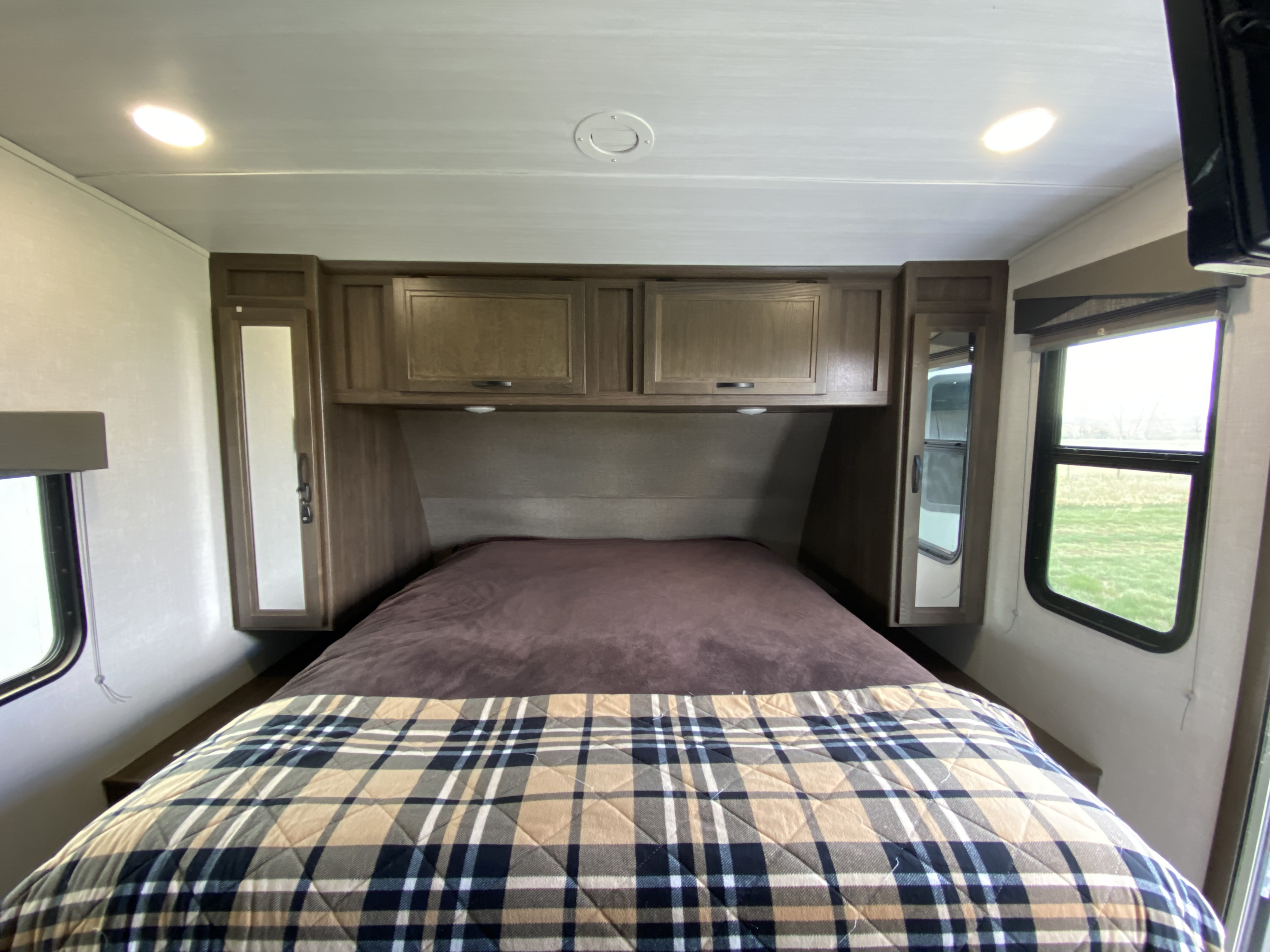 Cozy RV rental bedroom features a comfortable bed with plaid comforter, overhead storage, and natural light from windows.. Winnebago Winnebago 2021