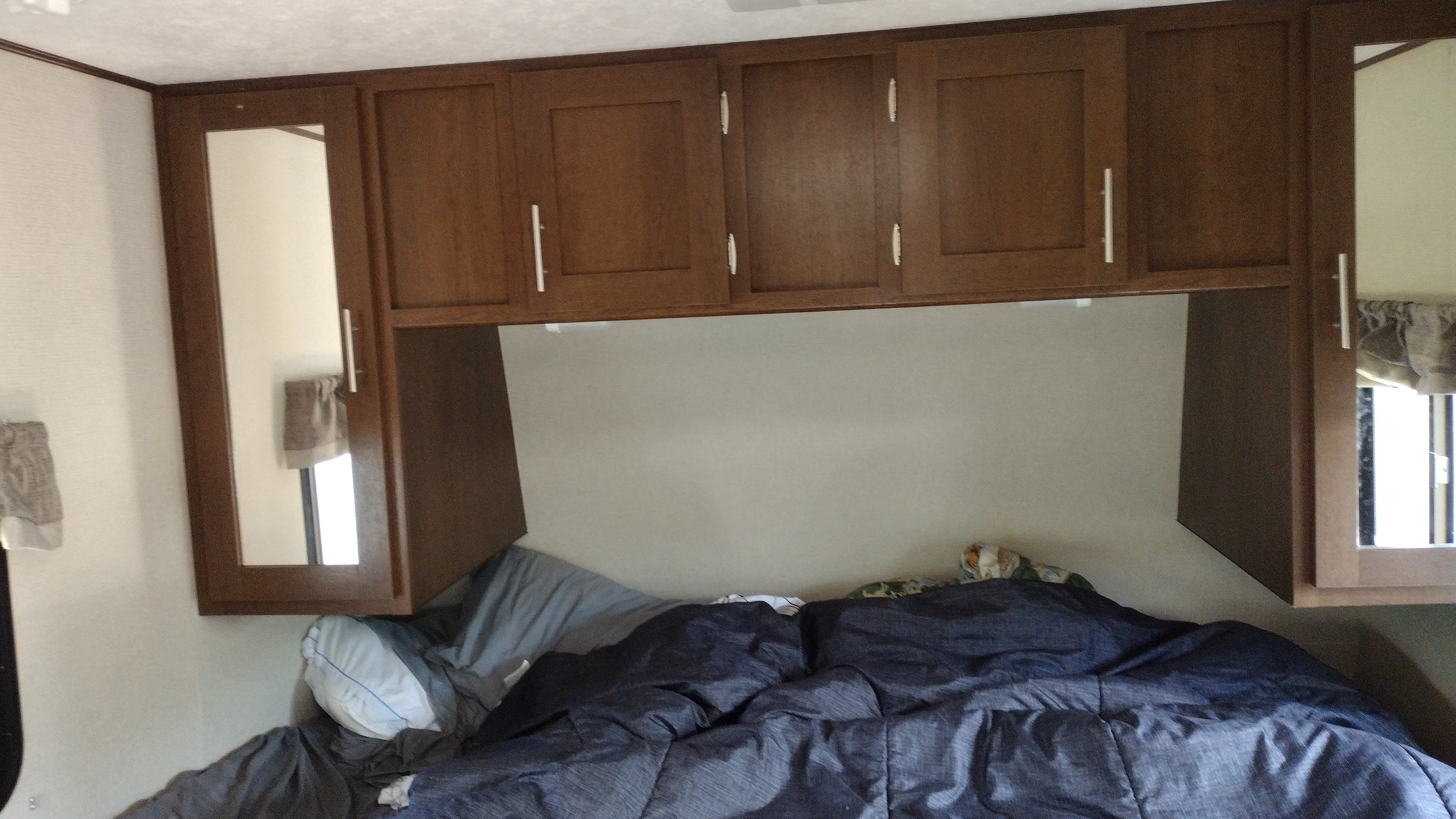 Inside an RV rental bedroom with wooden overhead cabinets, mirrored side cabinets, and a comfy bed with blue bedding.. Avenger ATI 2019
