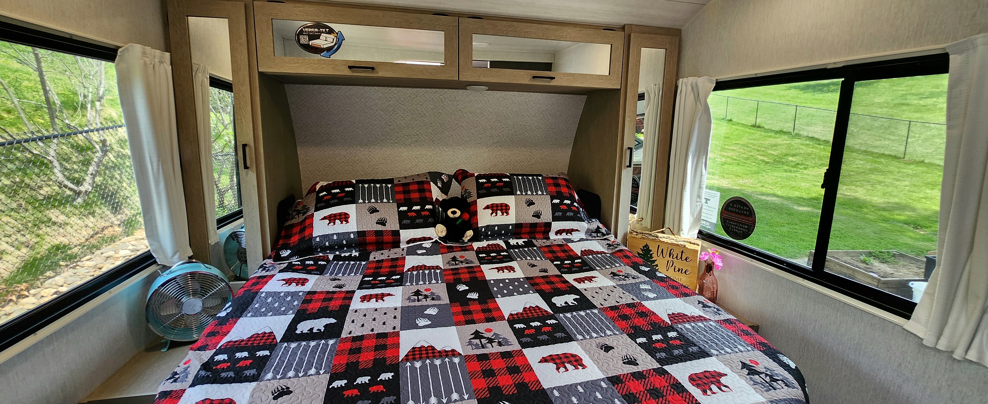 Cozy RV rental bedroom with a bear-themed quilt, overhead storage, and large windows overlooking green landscapes.. Forest River Wildwood Platinum 2024