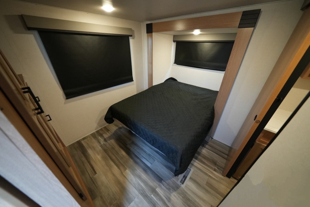 An RV rental's compact bedroom features a black-quilted bed, two windows with blinds, and light wood decor.. Prime Time Sanibel 2023