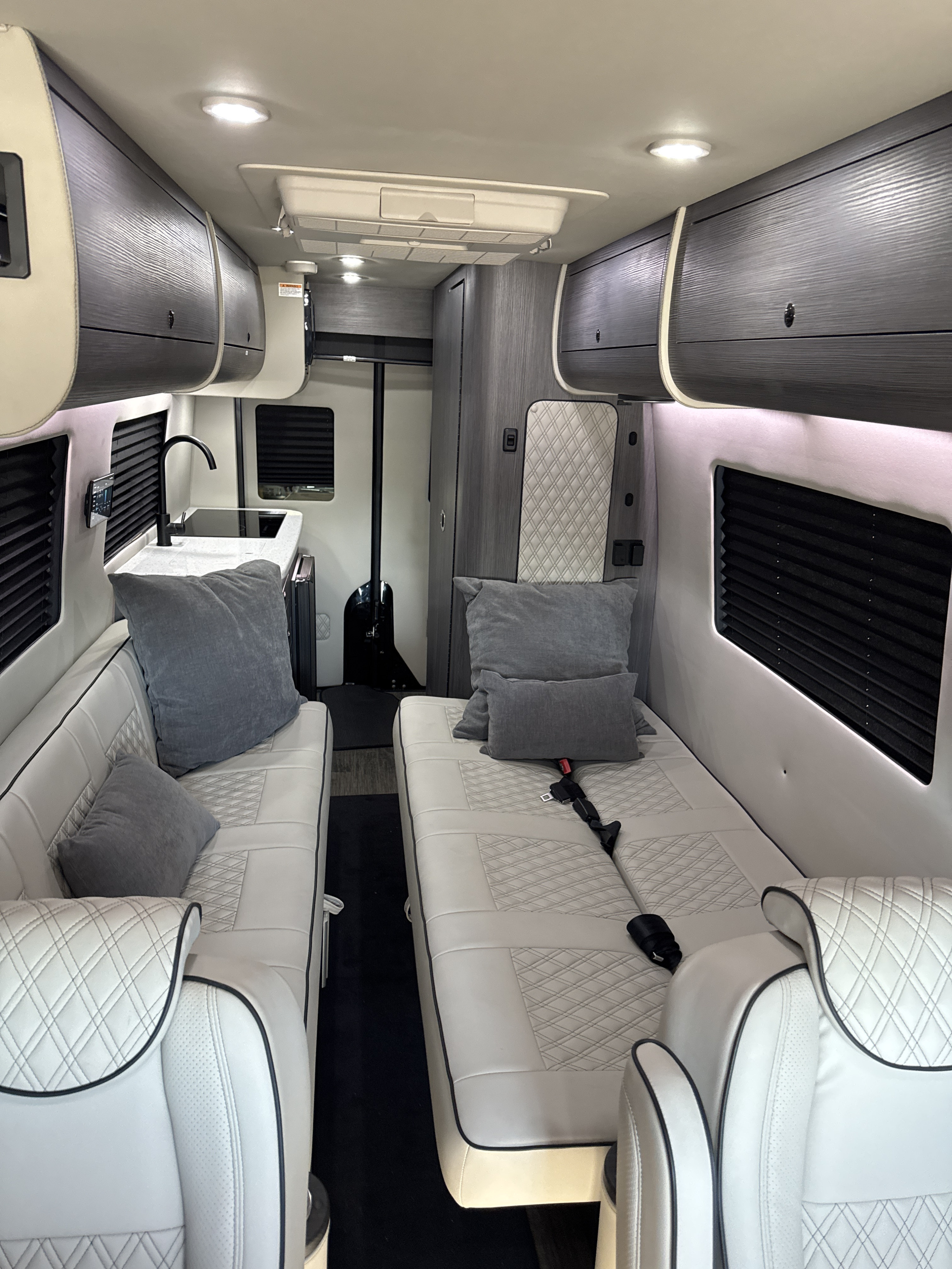 Bright, modern RV rental interior with white quilted seating, compact kitchen, and overhead storage for comfortable travel.. OGV luxury sprinter V-TOUR 2025
