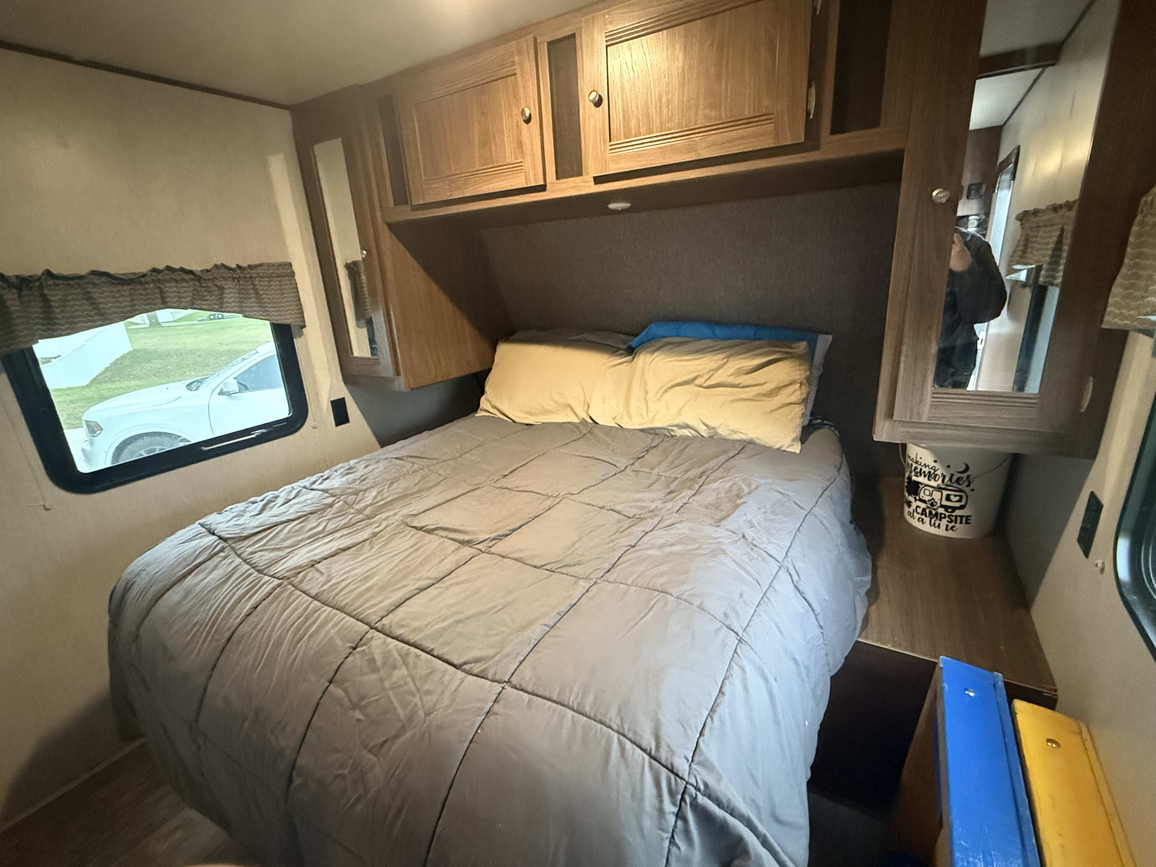 This cozy RV rental bedroom features a grey-covered bed, overhead wood cabinets, and a window. Ideal for your next camper trip.. Heartland RVs Pioneer 2018