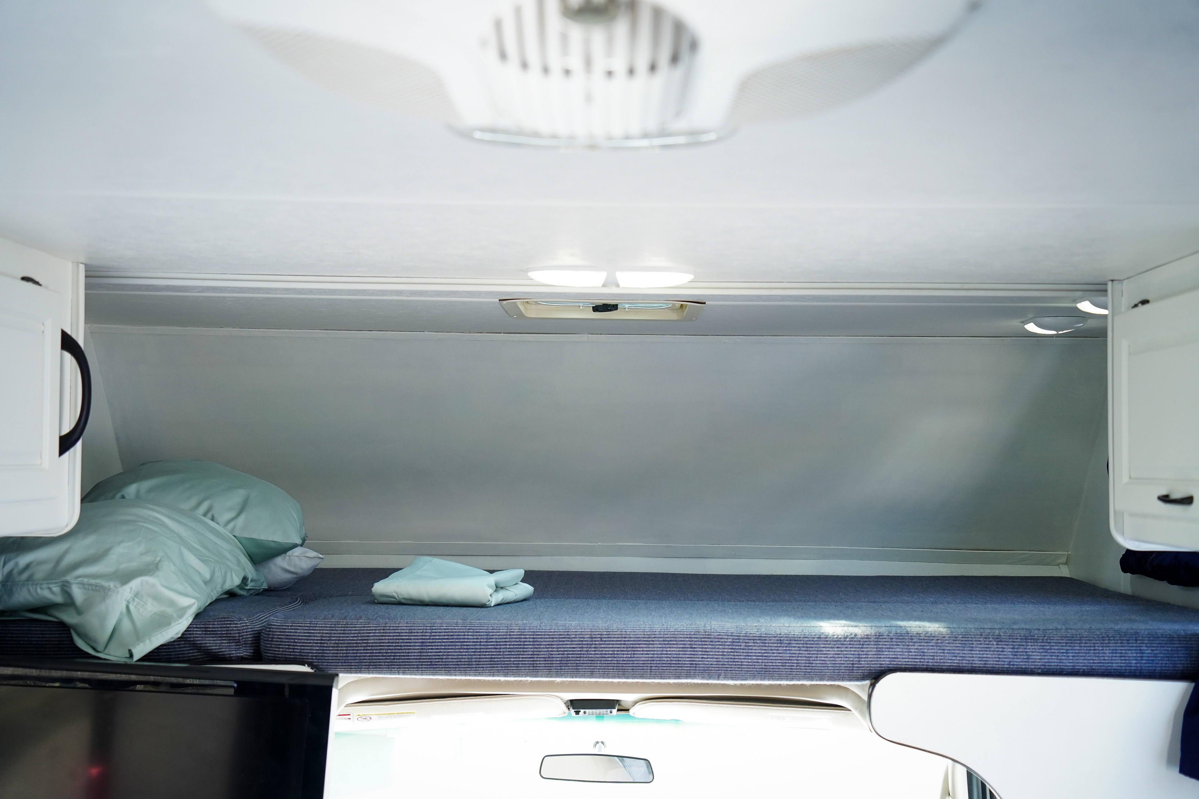 Spacious over-cab bed inside an RV rental, featuring blue bedding, pillows, and white storage cabinets. Brightly lit.. Thor Majestic 2014