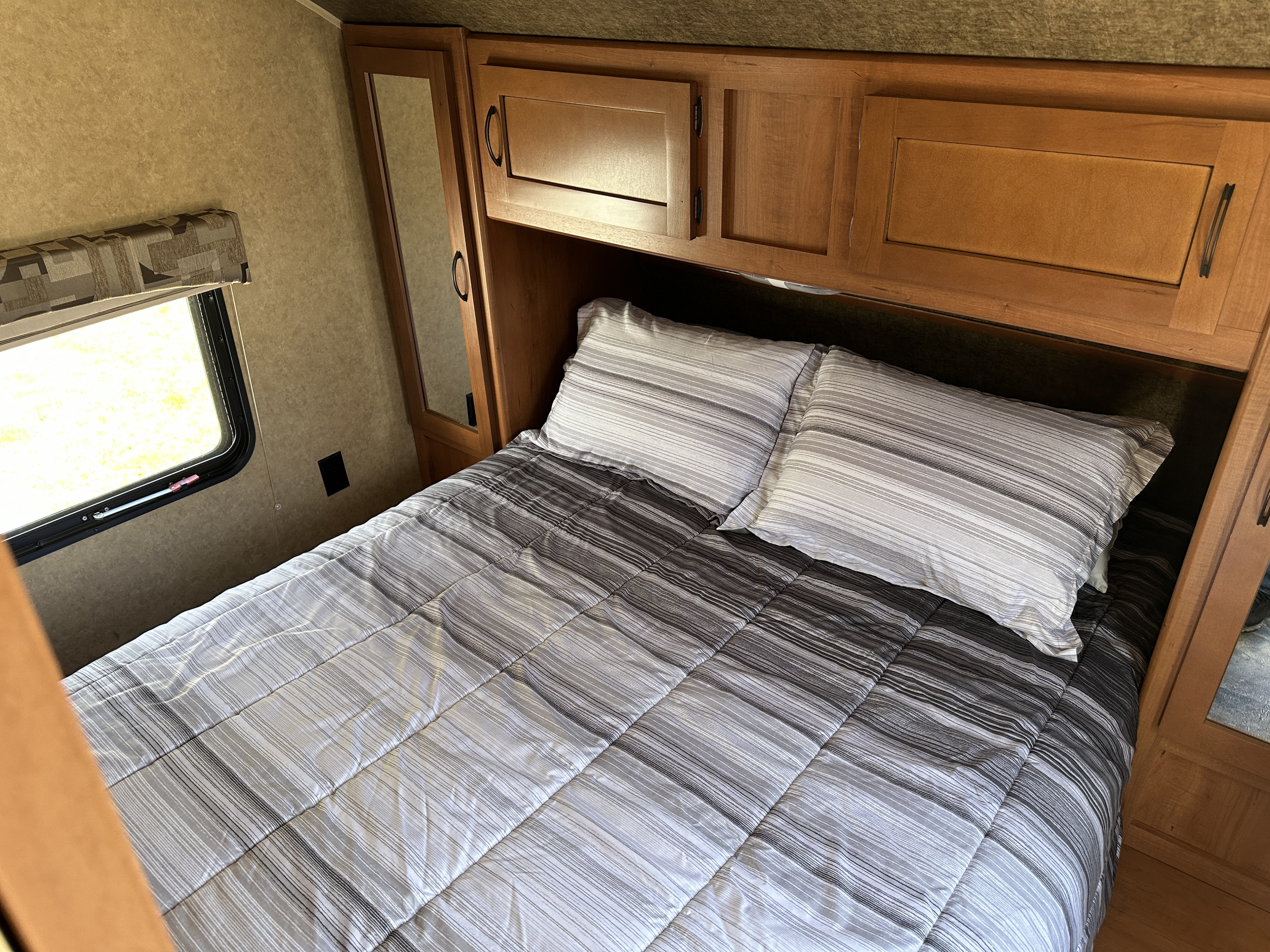Cozy RV rental bedroom featuring striped comforter, two pillows, overhead wood cabinets, and a window.. Forest River Cherokee 2015