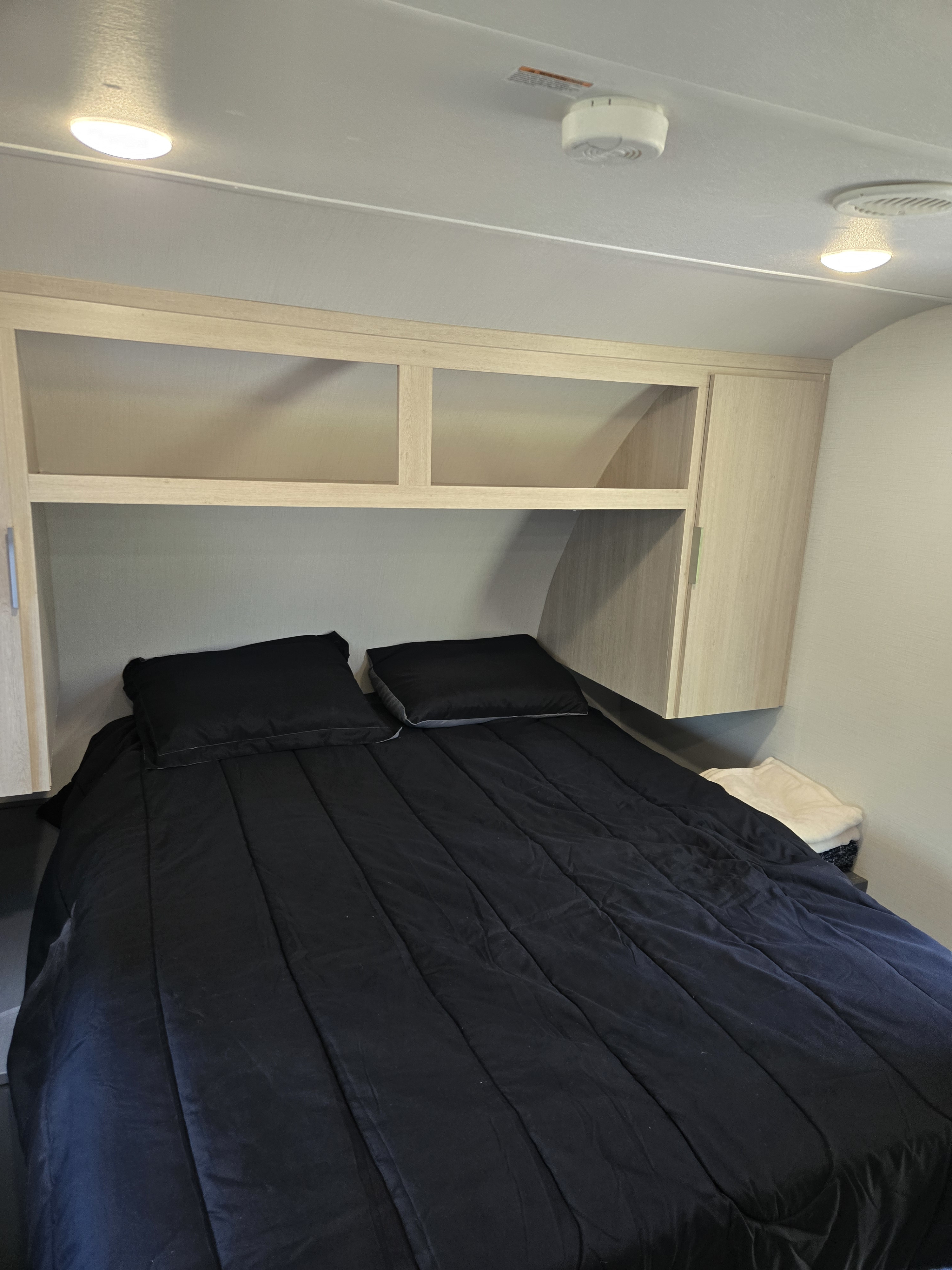 RV rental bedroom with black bedding, light wood overhead storage, and a smoke detector on the ceiling.. Prime Time Tracer 2024