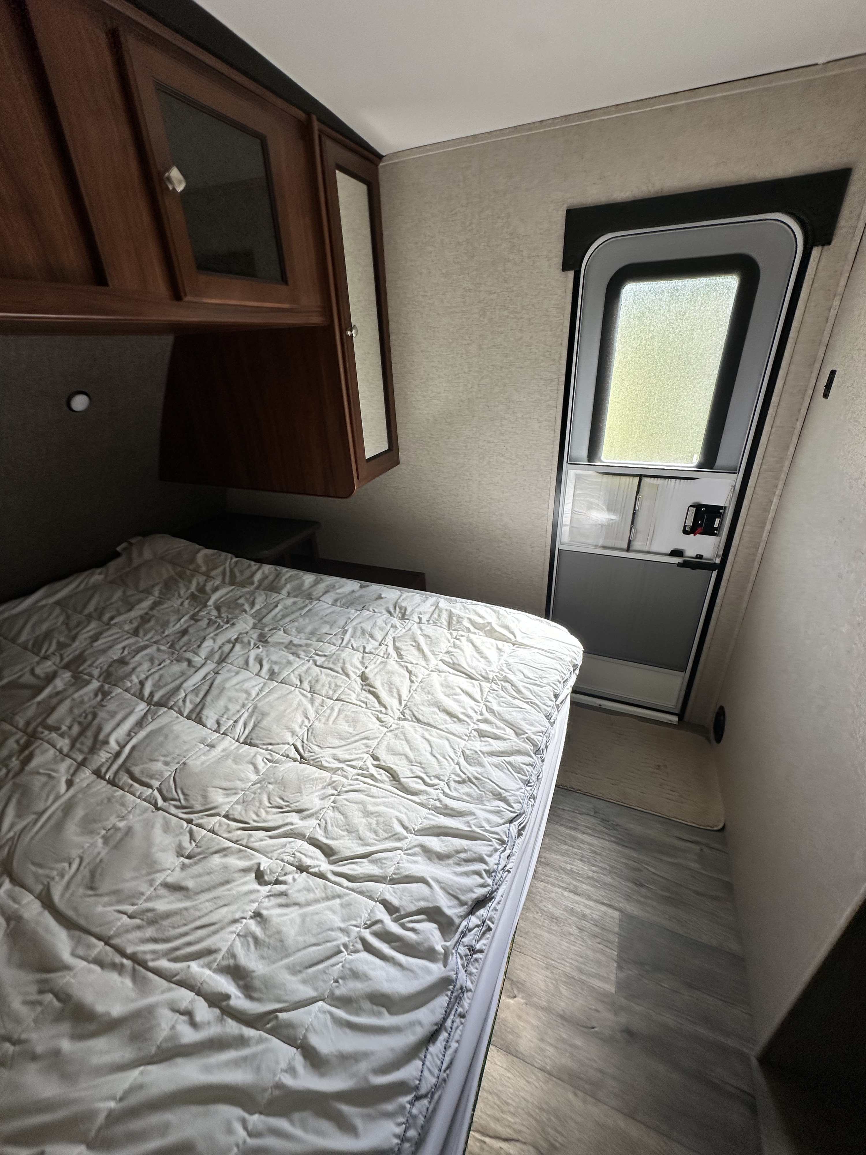 A cozy RV rental bedroom interior. The bed with a quilted comforter dominates, with wood cabinets overhead and an entry door on the right.. Heartland RVs Mallard 2017