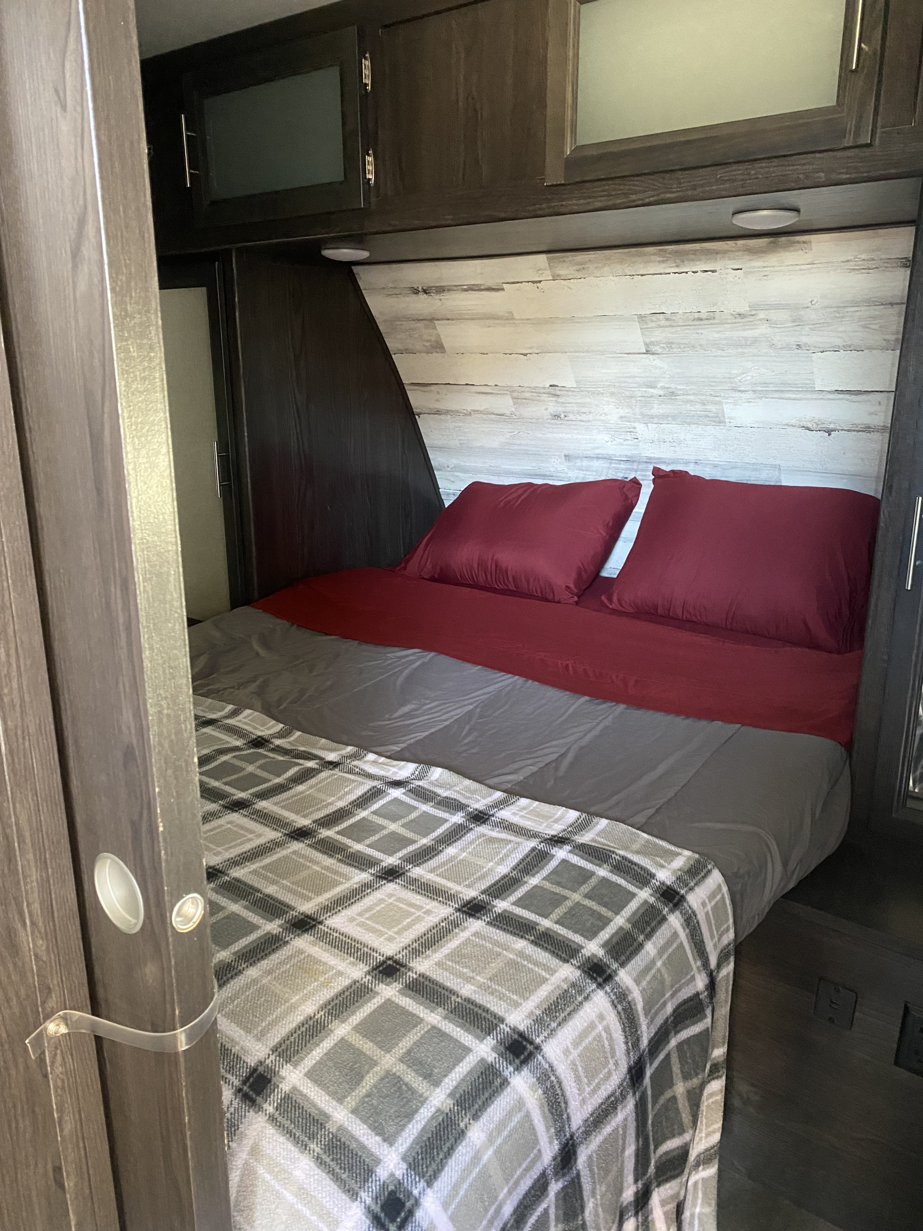 Cozy RV rental bedroom featuring a comfortable bed with red pillows, gray comforter, and plaid blanket. Overhead cabinets are visible.. Dutchmen Kodiak Ultra Lite 2020