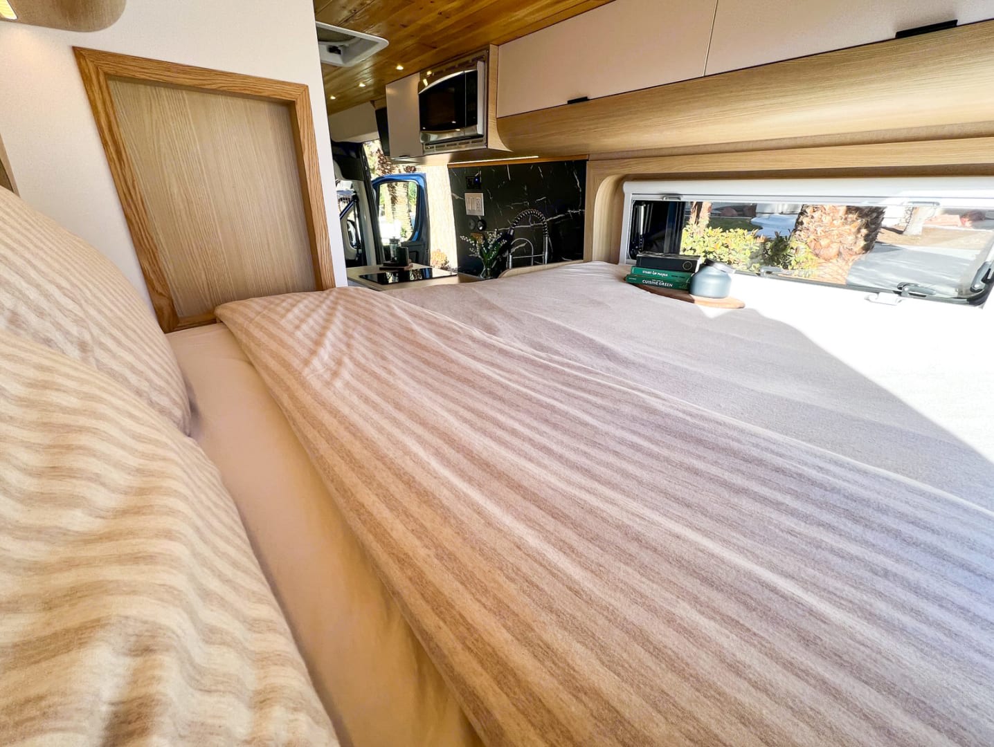 Cozy RV rental bedroom with striped bedding, a window view, and a compact kitchen visible in the background.. Dodge Ram Promaster 3500 2022