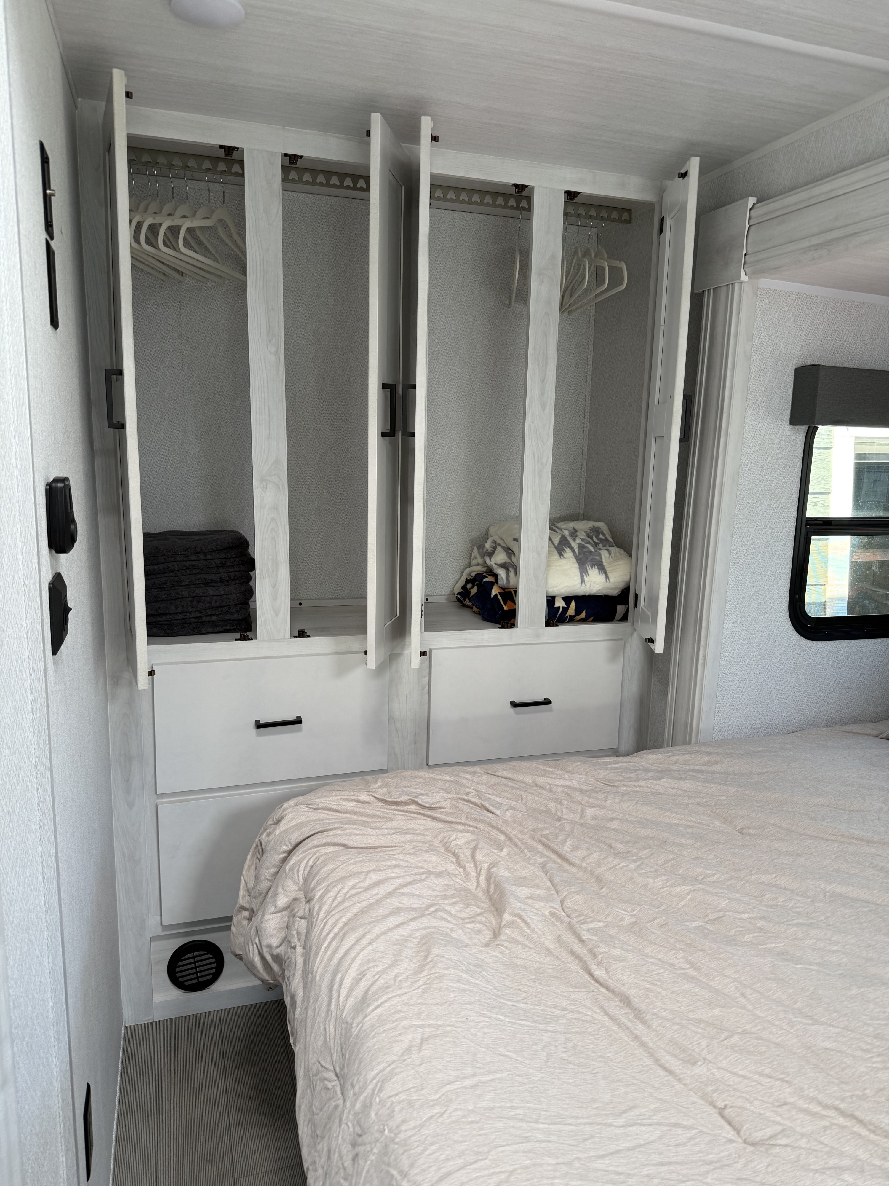 RV rental bedroom with light-toned wardrobe: open doors reveal hangers, folded towels, and blankets, plus lower drawers.. East to West Entrada M-Class 2025