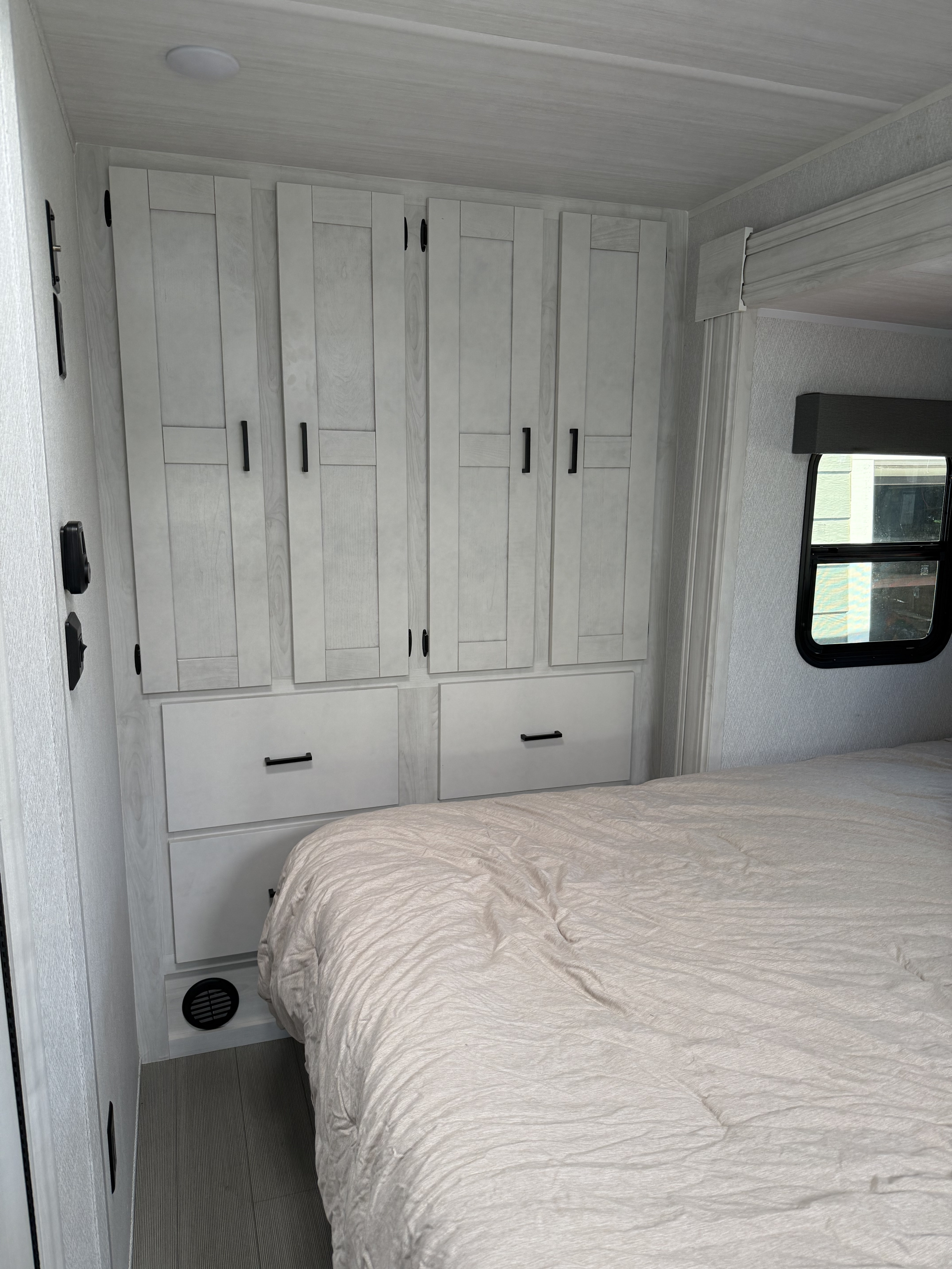 RV rental bedroom with light-colored bed, built-in white cabinets, drawers, and a small side window for optimized storage and comfort.. East to West Entrada M-Class 2025