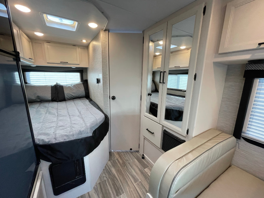 Inside an rv rental: a cozy bedroom with gray bedding, mirrored wardrobe, and seating next to a window.. Thor Freedom Elite 2022