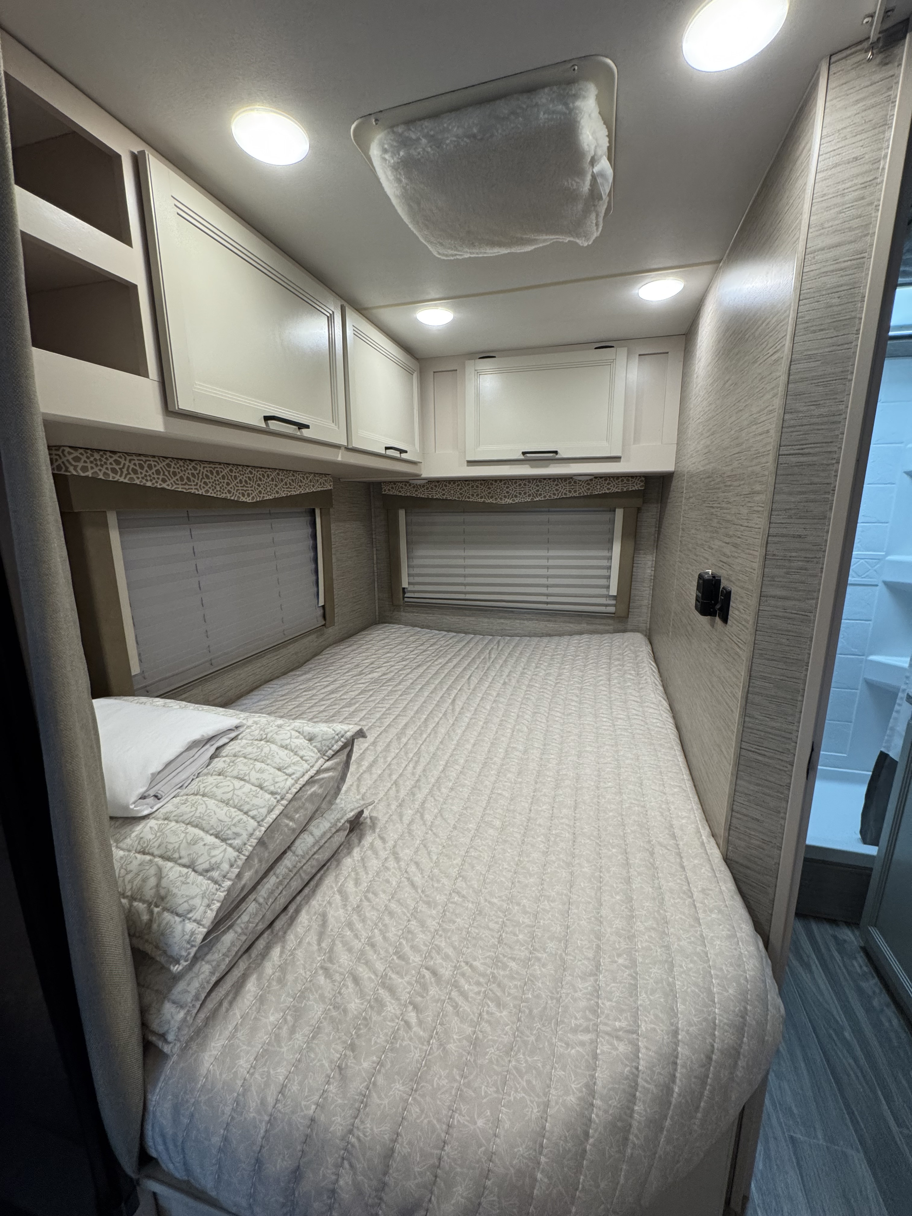 Inside an RV rental, a compact bedroom with a light gray quilted bed, overhead cabinets, and windows with blinds.. Thor Freedom Elite 2022