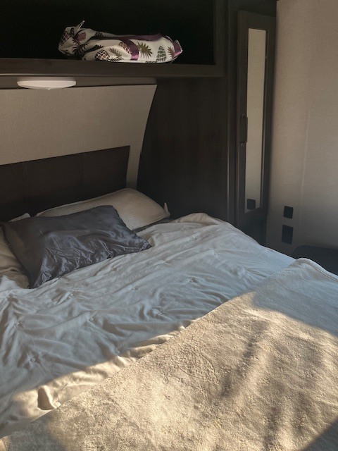 RV rental bedroom: A cozy bed with neutral bedding, overhead light, and mirrored storage cabinet are ready for your trip.. Grand Design Transcend Xplor 2023
