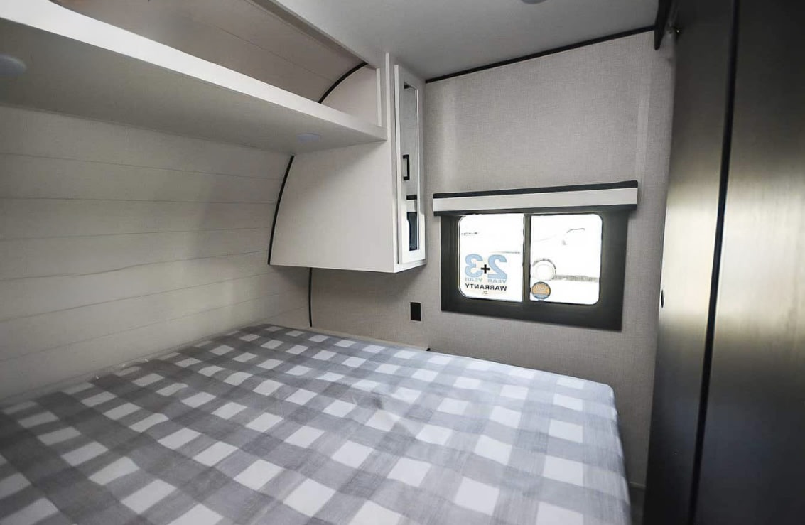 RV rental bedroom: cozy bed with gray checker pattern, overhead storage, window. A functional and compact interior.. Jayco Jay Flight 2024
