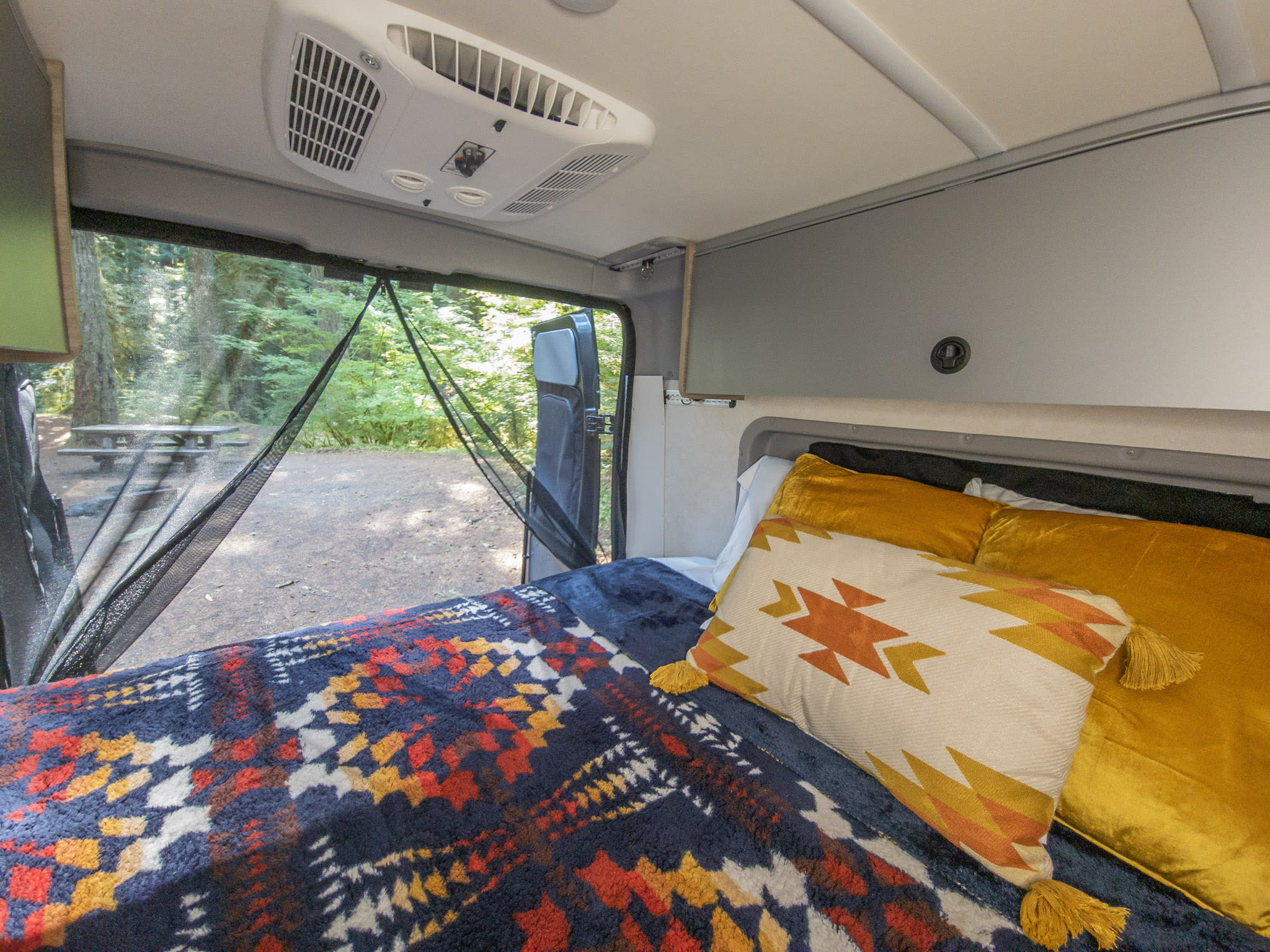 Comfortable RV rental interior with a cozy bed and AC, offering a screened view of a peaceful forest campsite.. Winnebago Solis 2023