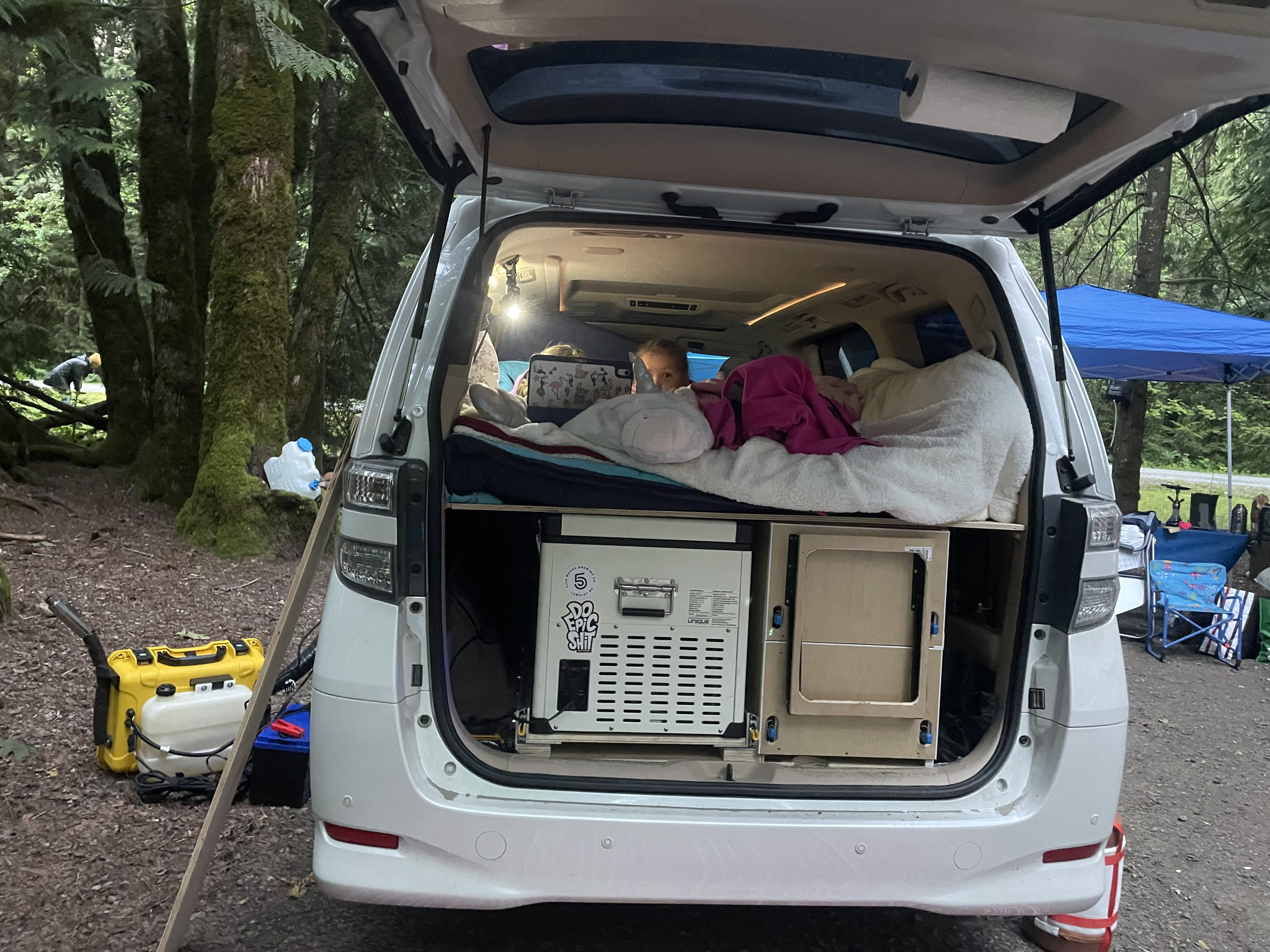 Custom minivan setup as an RV rental alternative for camping. A child is nestled in the back bed, with fridge & power below. Forest camp.. Toyota Dreamseeker Voyager 2008