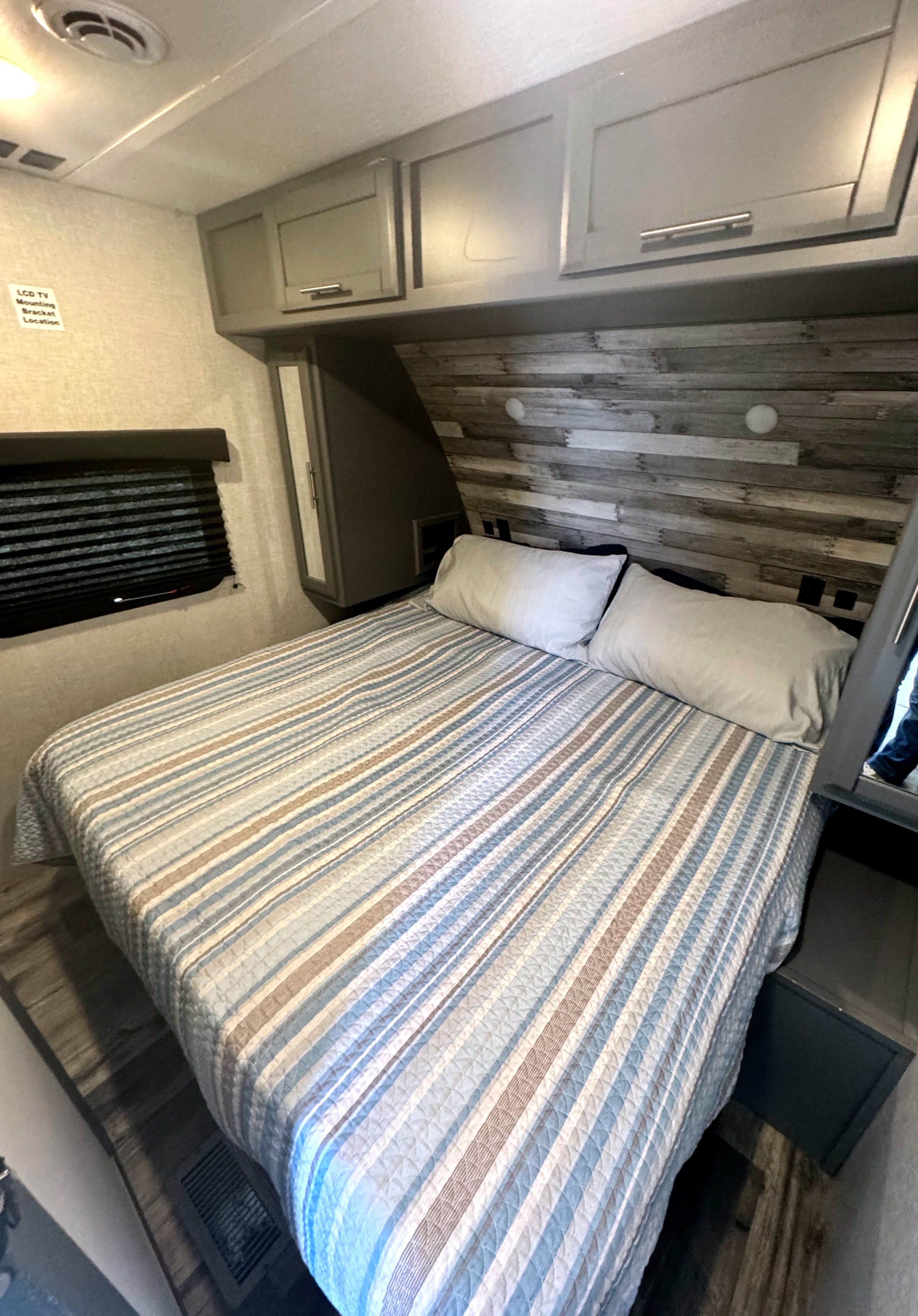 This RV rental's cozy bedroom features a striped bed, wood-paneled wall, overhead cabinets, and a window with blinds.. Crossroads Volante 2022