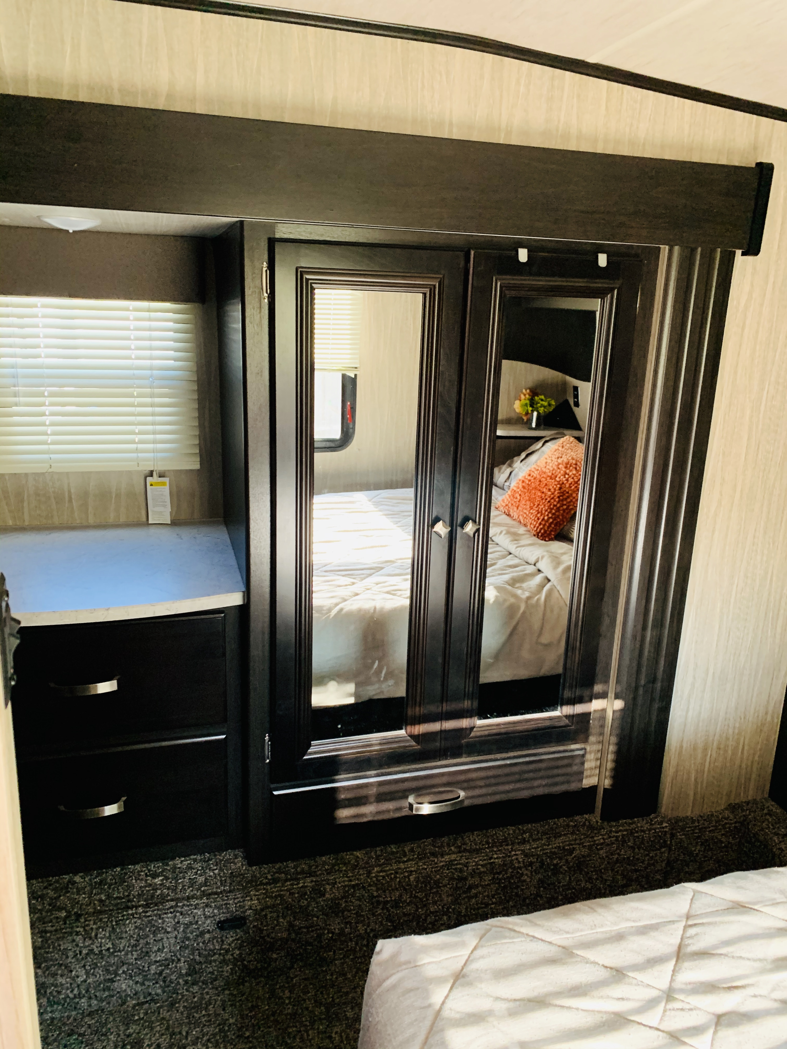 View of an RV rental bedroom with a mirrored closet, dark wood cabinetry, window with blinds, and comfy bed reflection.. Heartland RVs Fuel Toy Hauler 2019
