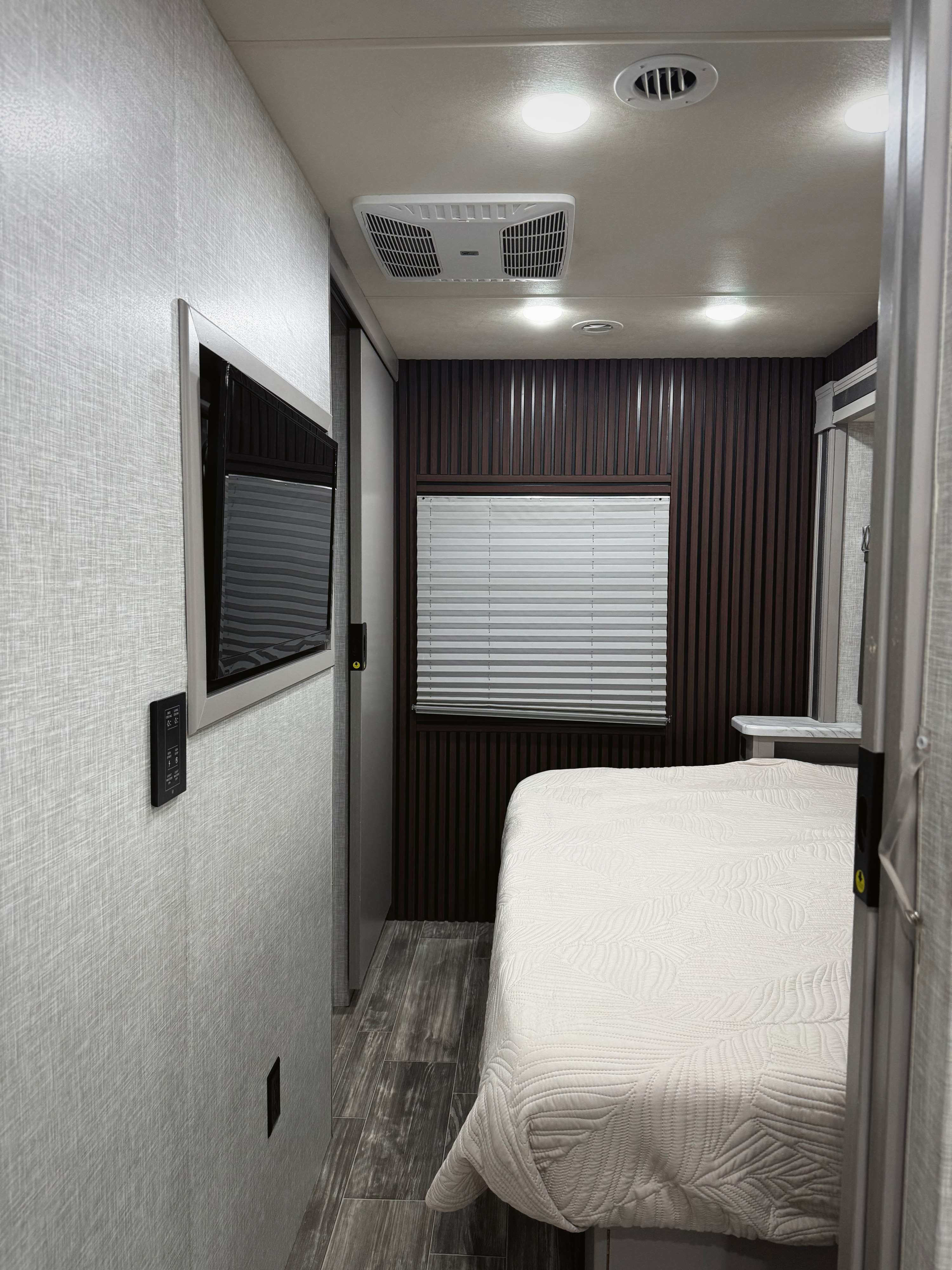 This RV rental bedroom features a comfortable bed, wall-mounted TV, light-textured walls, dark wood paneling, and ample lighting.. Thor Motor Coach Hurricane 2021