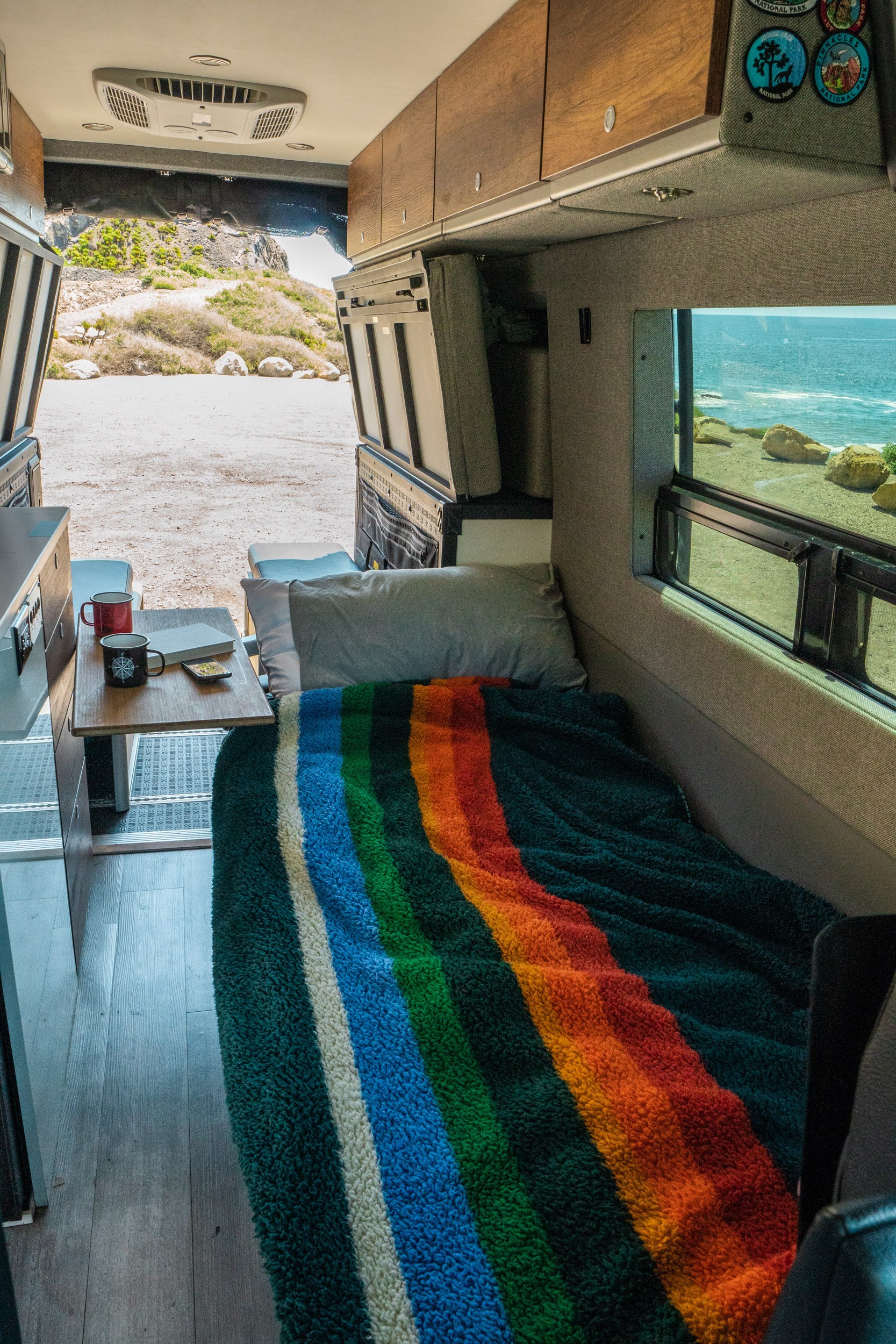 Cozy RV rental interior with bed, mugs, and a view of the ocean & landscape.. Storyteller Overland Storyteller Overland Class B 2022