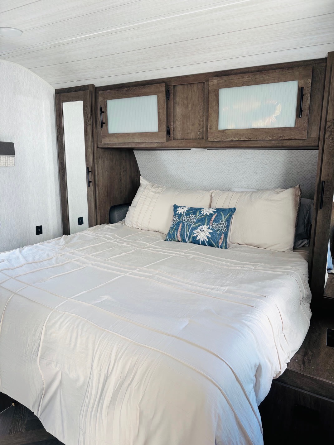 An RV rental bedroom: a large bed with light bedding, a blue accent pillow, and wood cabinets above.. Forest River Wildwood 2022