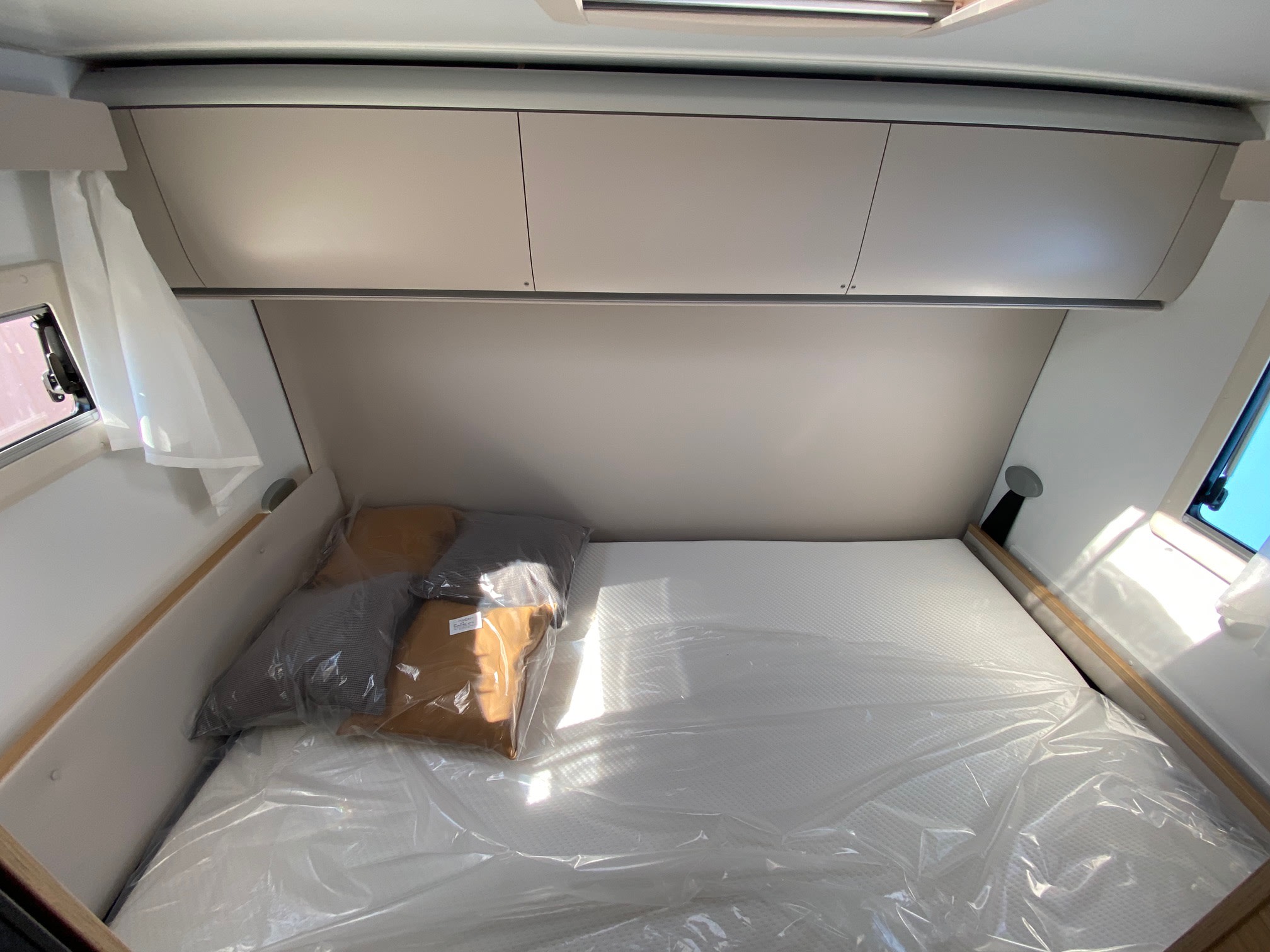 Clean RV rental bedroom, featuring a new, plastic-wrapped mattress and pillows, overhead cabinets, and bright windows.. Adria Coral XL Axess 600 DP 2025