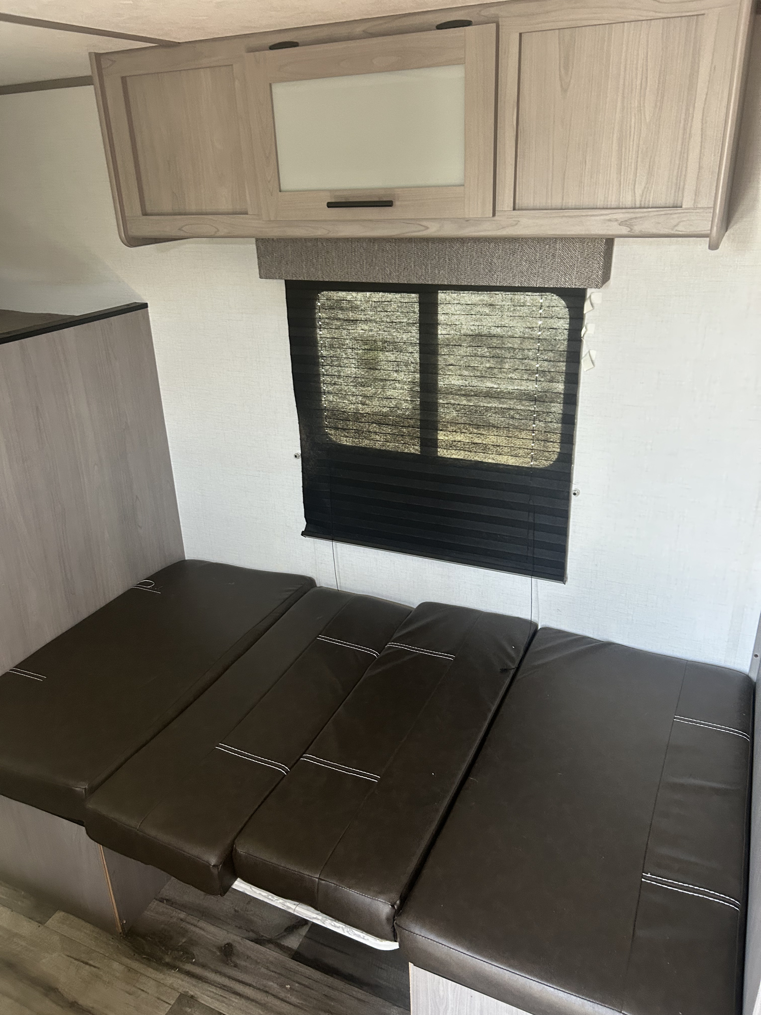 Inside an RV rental: light wood cabinets, window with blinds, and dark brown cushions for a dinette that converts to a bed.. Dutchmen Coleman 17B 2022