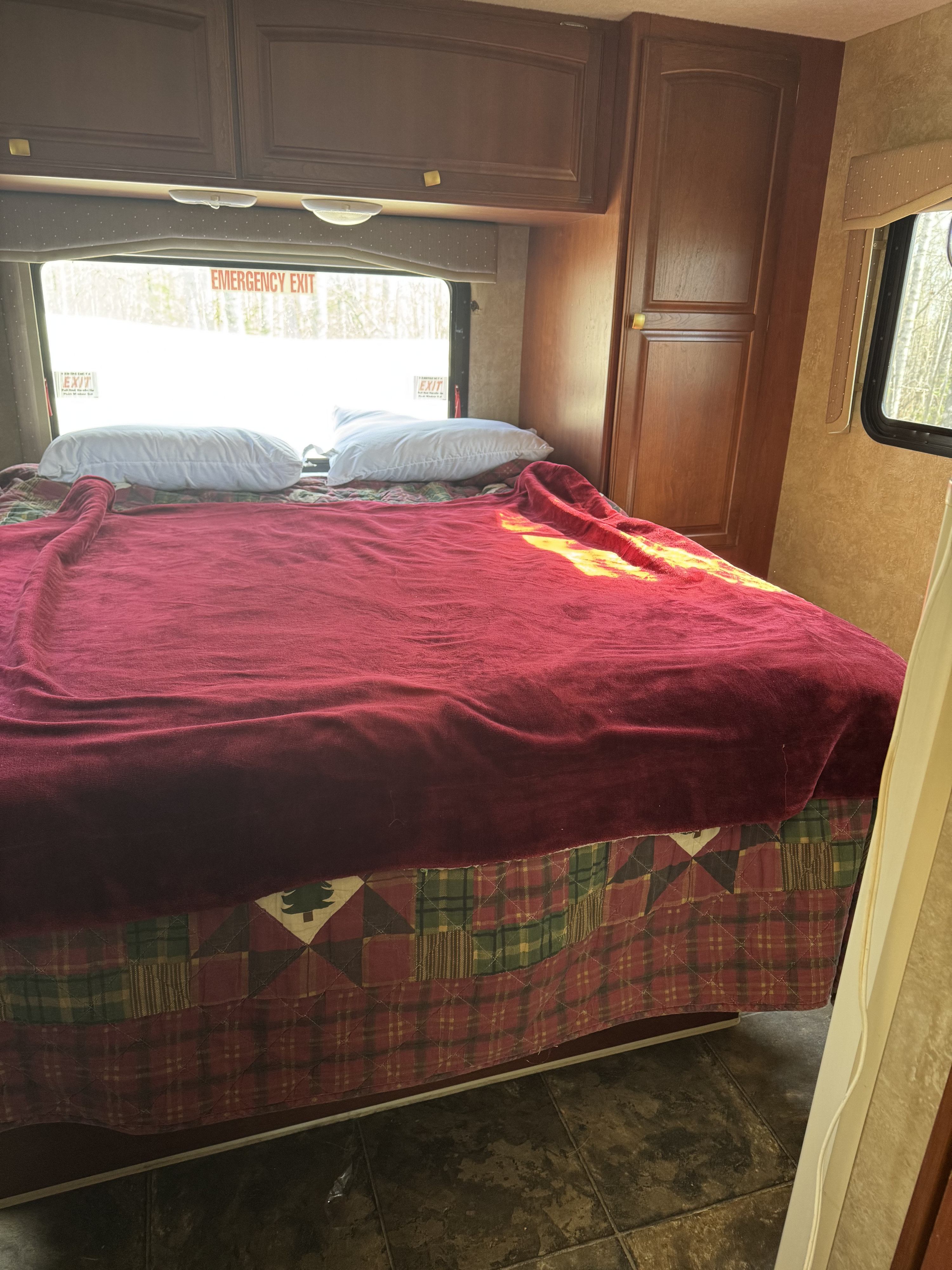 Inside a cozy RV rental: a bed with red blanket, plaid skirt, and pillows, near a window showing snowy trees.. Winnebago Chalet 2014