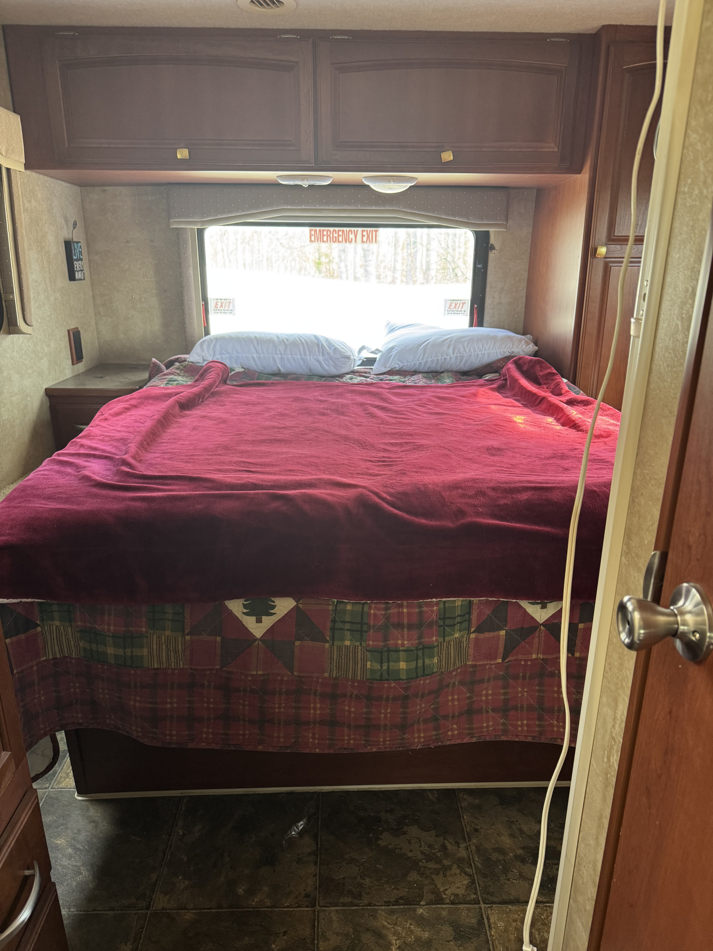 RV rental bedroom with a comfy bed, red blanket, overhead cabinets, and an emergency exit window.. Winnebago Chalet 2014