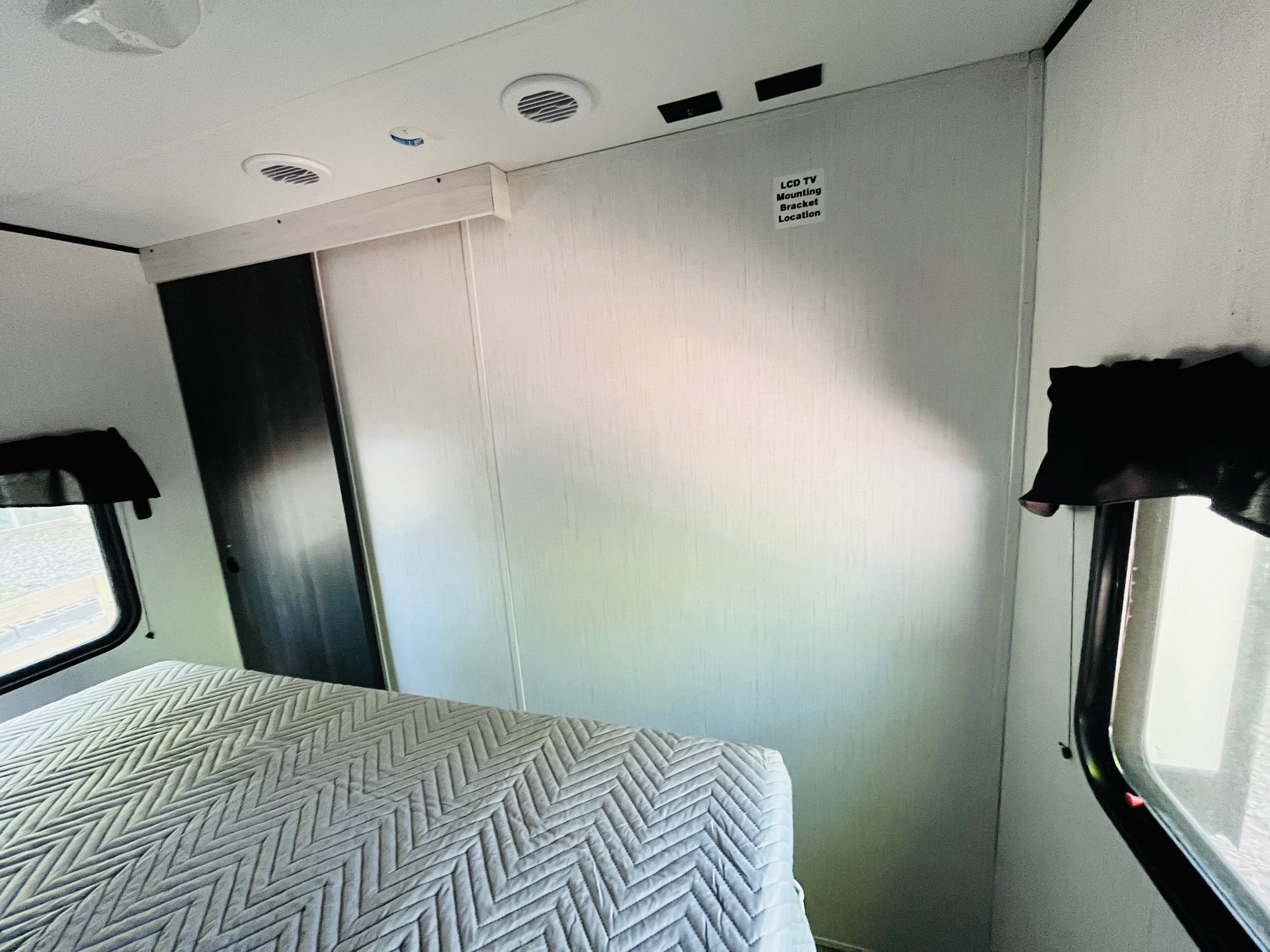 Inside an RV rental: a bright bedroom with a quilted bed, windows, and a dedicated wall for a TV mount.. Heartland RVs Trail Runner 2023