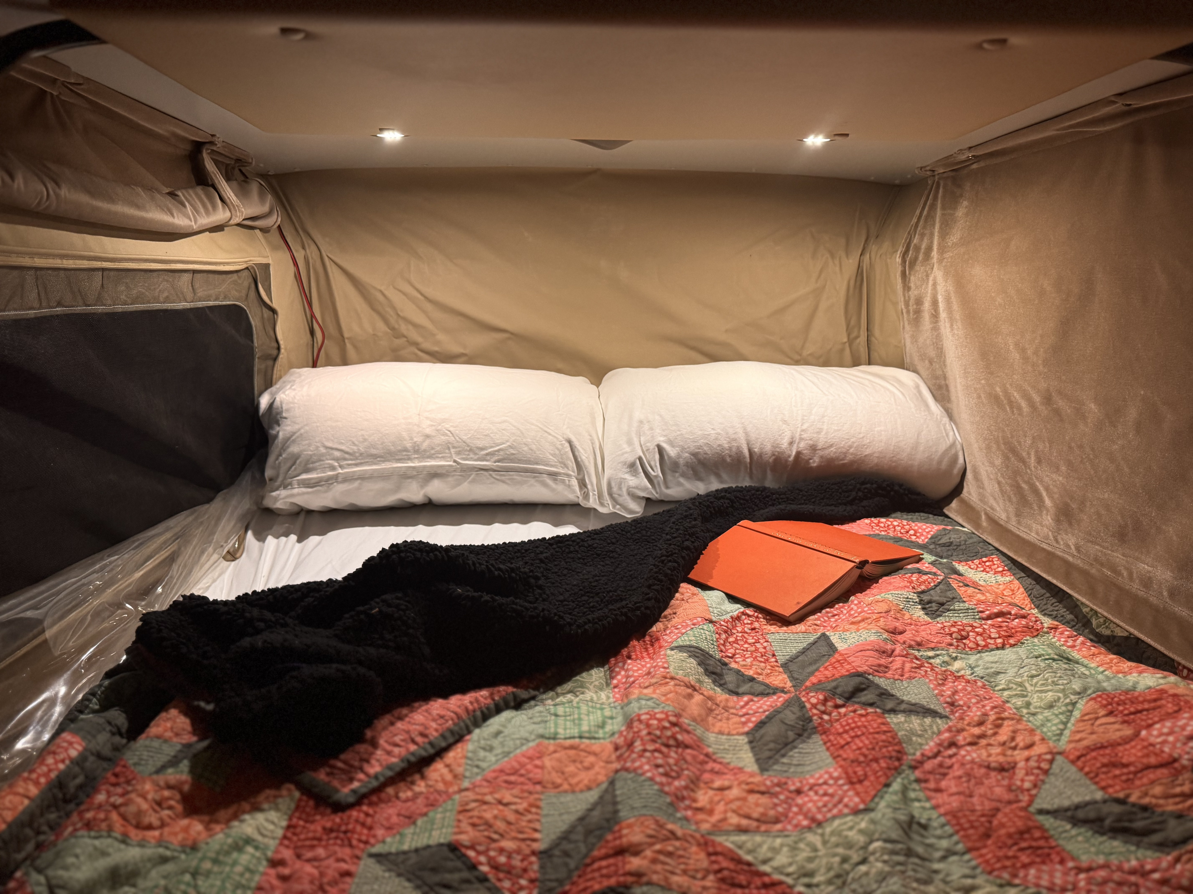 Cozy RV rental bed setup with white pillows, a colorful quilt, and an open journal, ideal for a relaxing night.. Sportsmobile Regular Body 2013