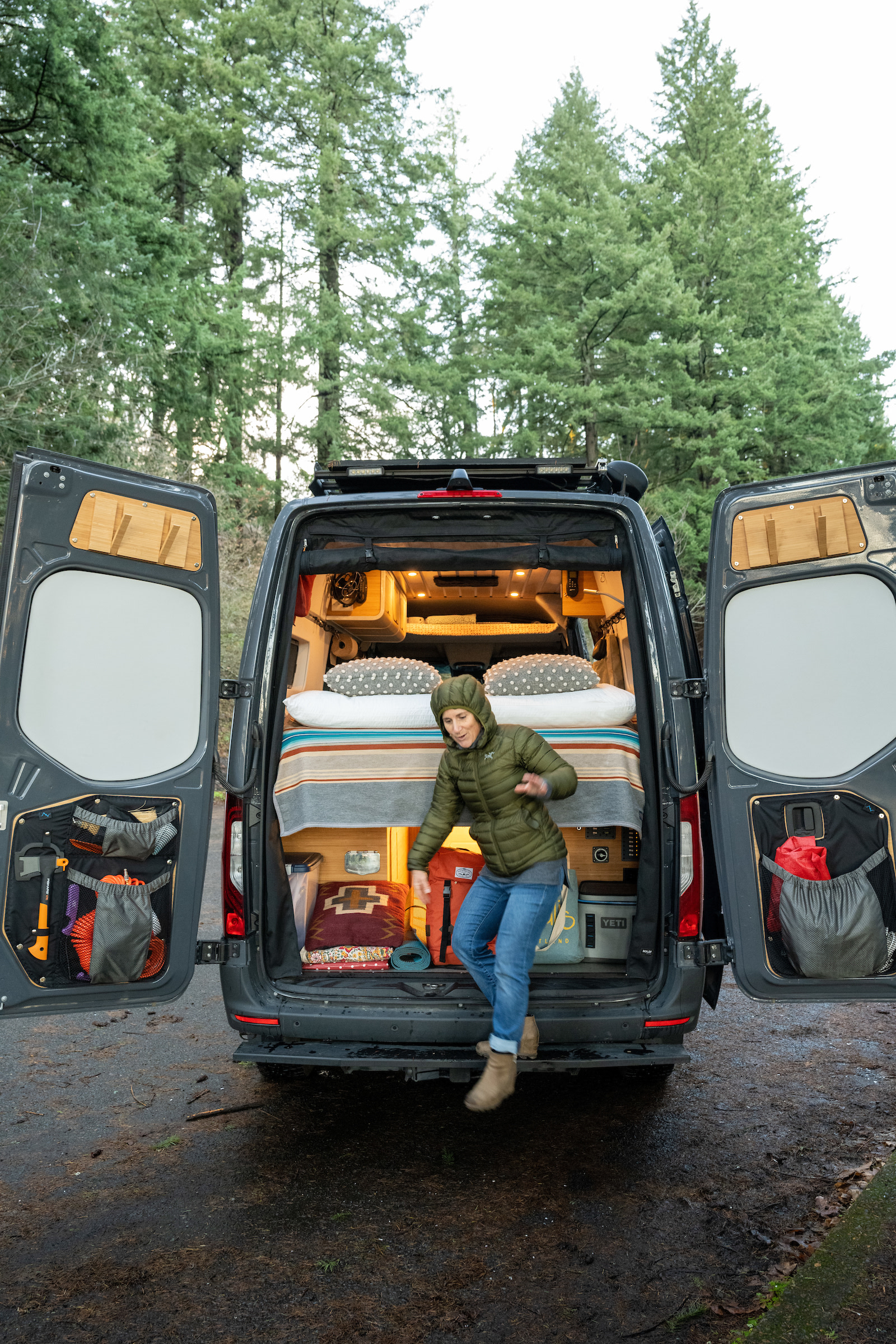 This custom camper van for RV rental allows easy outdoor access. A woman steps out, revealing cozy interior & gear storage.. Mercedes 144 Sprinter Van Custom build by Overland Van Project Portland Oregon 2023