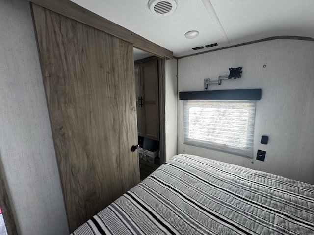 Cozy RV rental bedroom featuring a striped bed, window with blinds, TV mount, and a sliding barn door.. Forest River Cherokee Alpha Wolf 2021