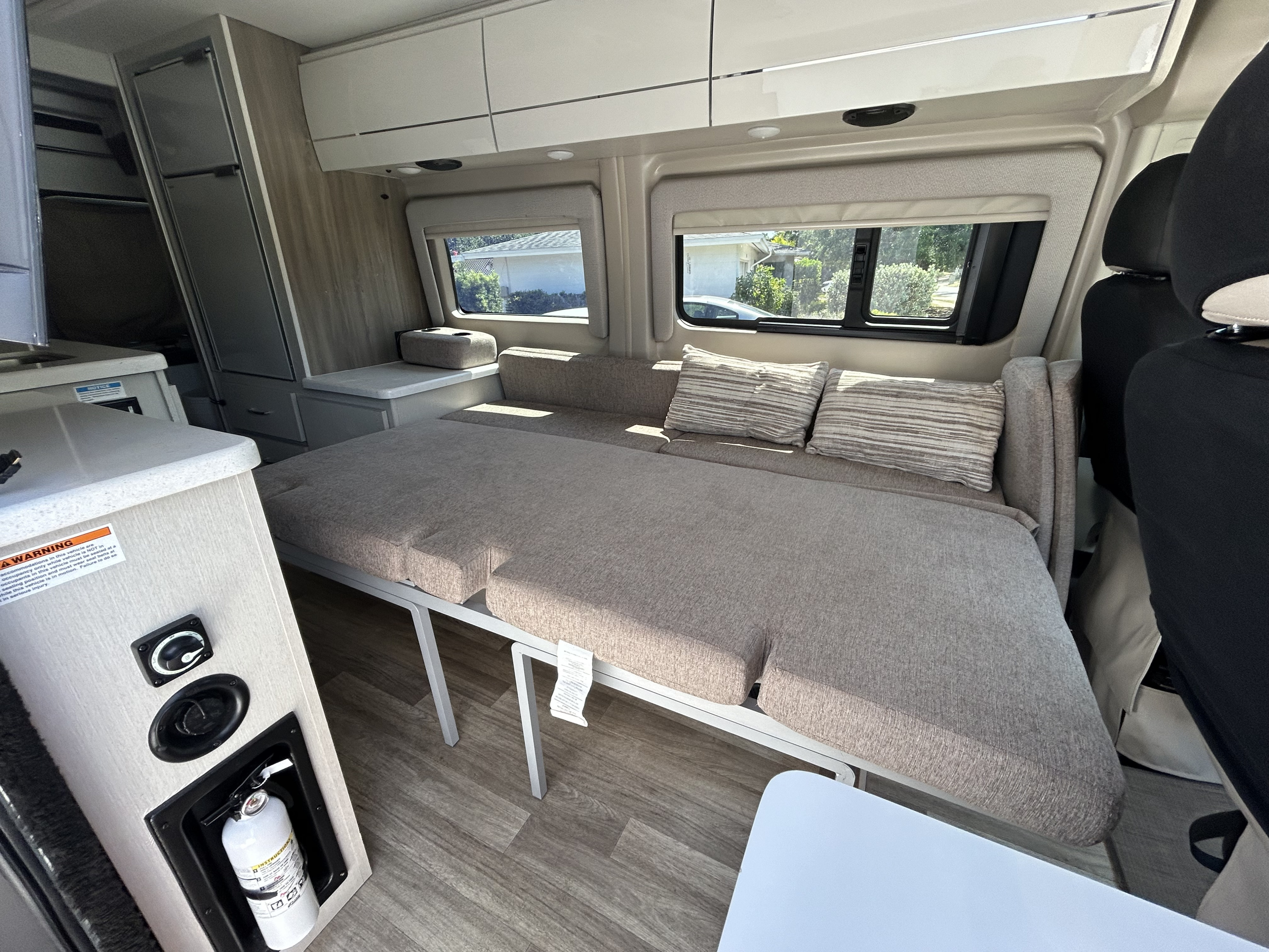 Compact RV rental interior featuring a convertible bed/seating area, windows, overhead storage, and a kitchenette.. Thor Tellaro 2023