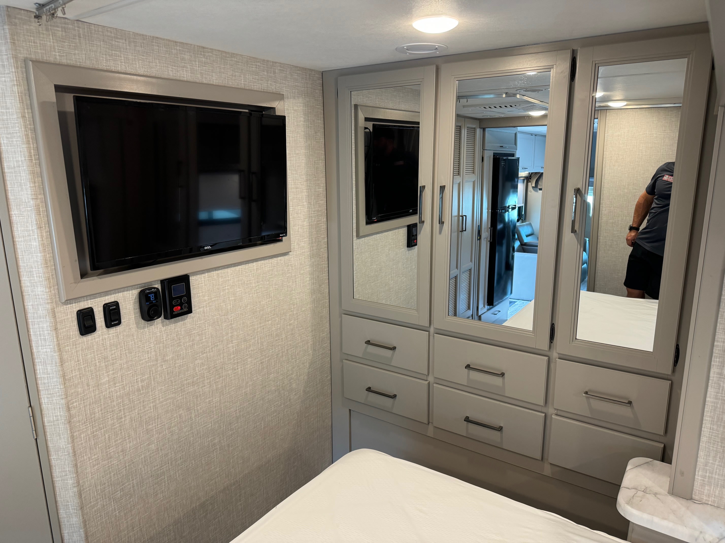 RV rental bedroom: wall-mounted TV, climate & slide-out controls, plus mirrored wardrobe with drawers. Compact comfort for travel.. Thor Motor Coach Geneva 2025