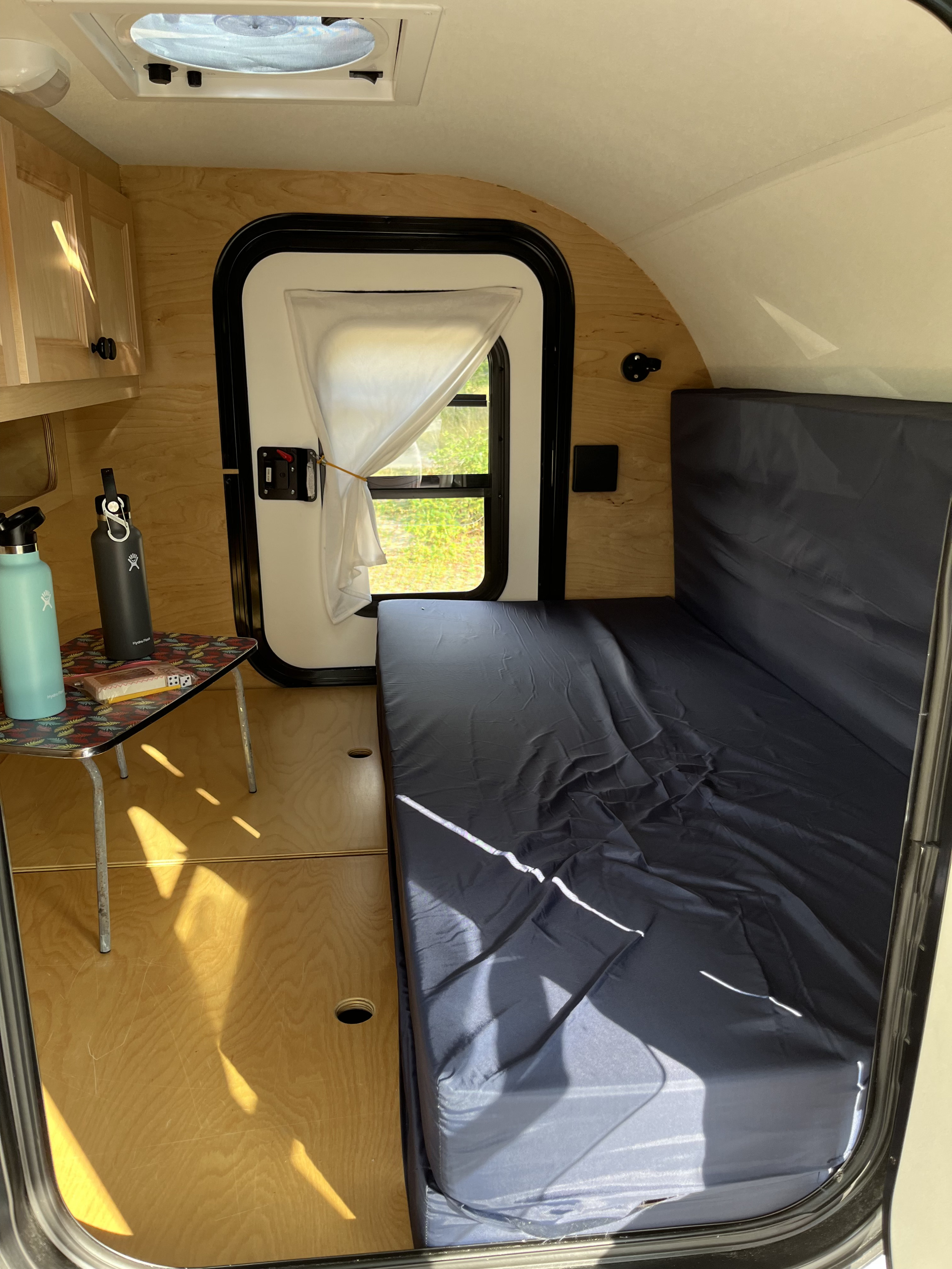 Inside an RV rental, a compact camper with a dark bed, wooden cabinets, and a small table holding water bottles.. Car-go Trailers Liberty2go XL 2021